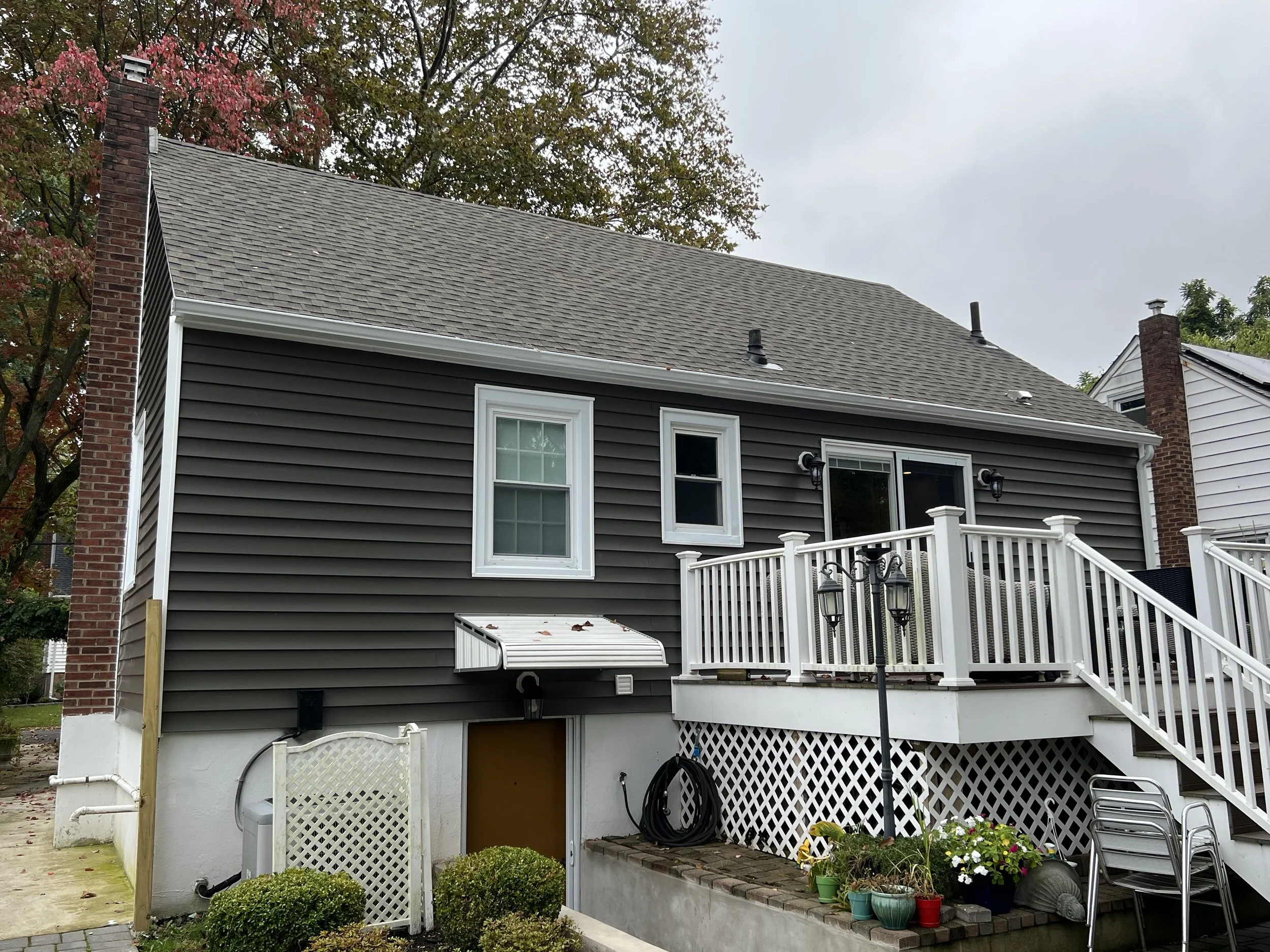 Rear view of a two-story house with gray siding, white trim, a wooden deck with railing, and three steps leading down. The house has two small windows on the upper level and a large sliding glass door on the deck. There are various potted plants, gar