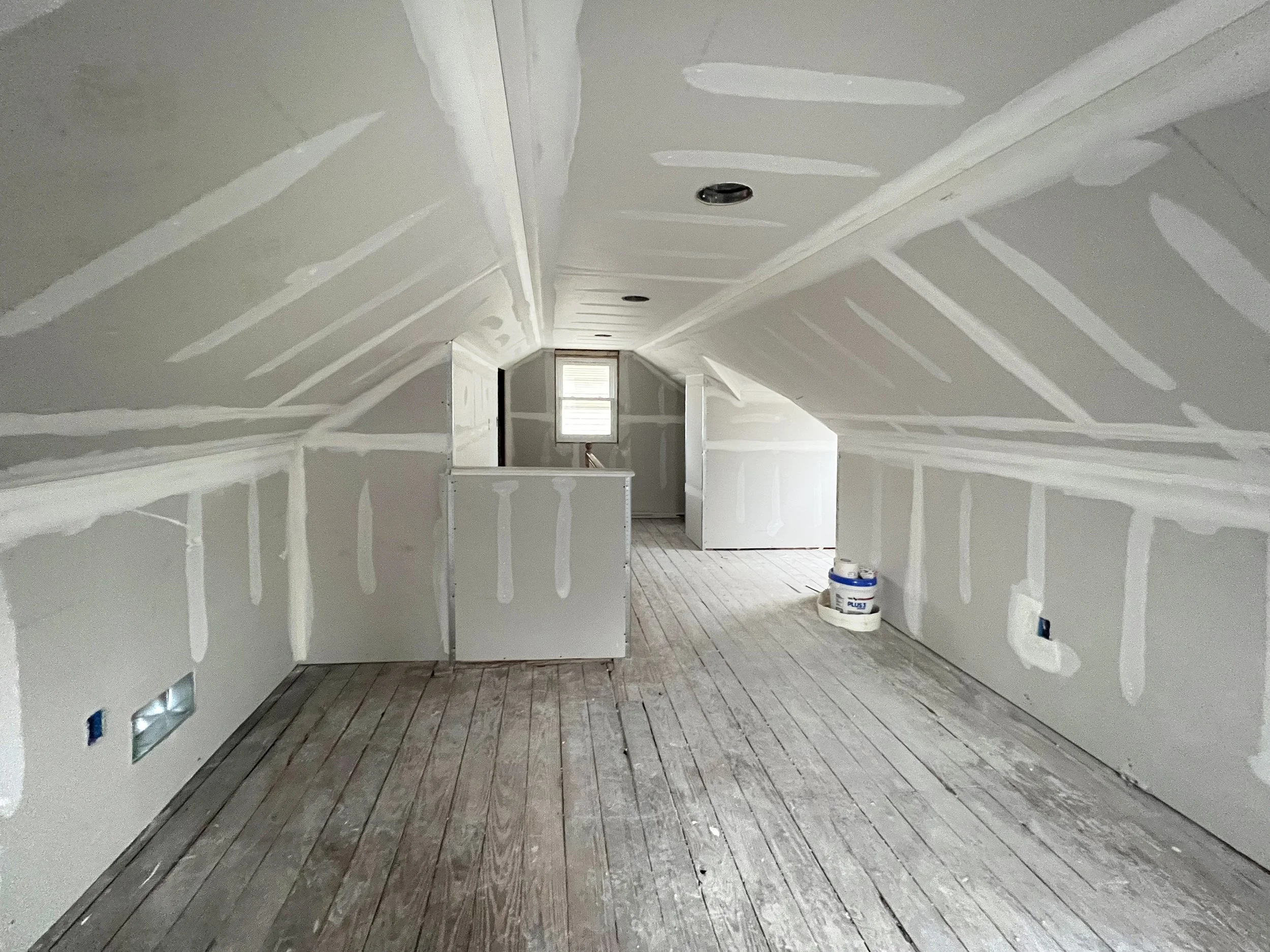 Attic room under construction with drywall installation, unfinished wooden floor, and electrical outlets and wires visible.