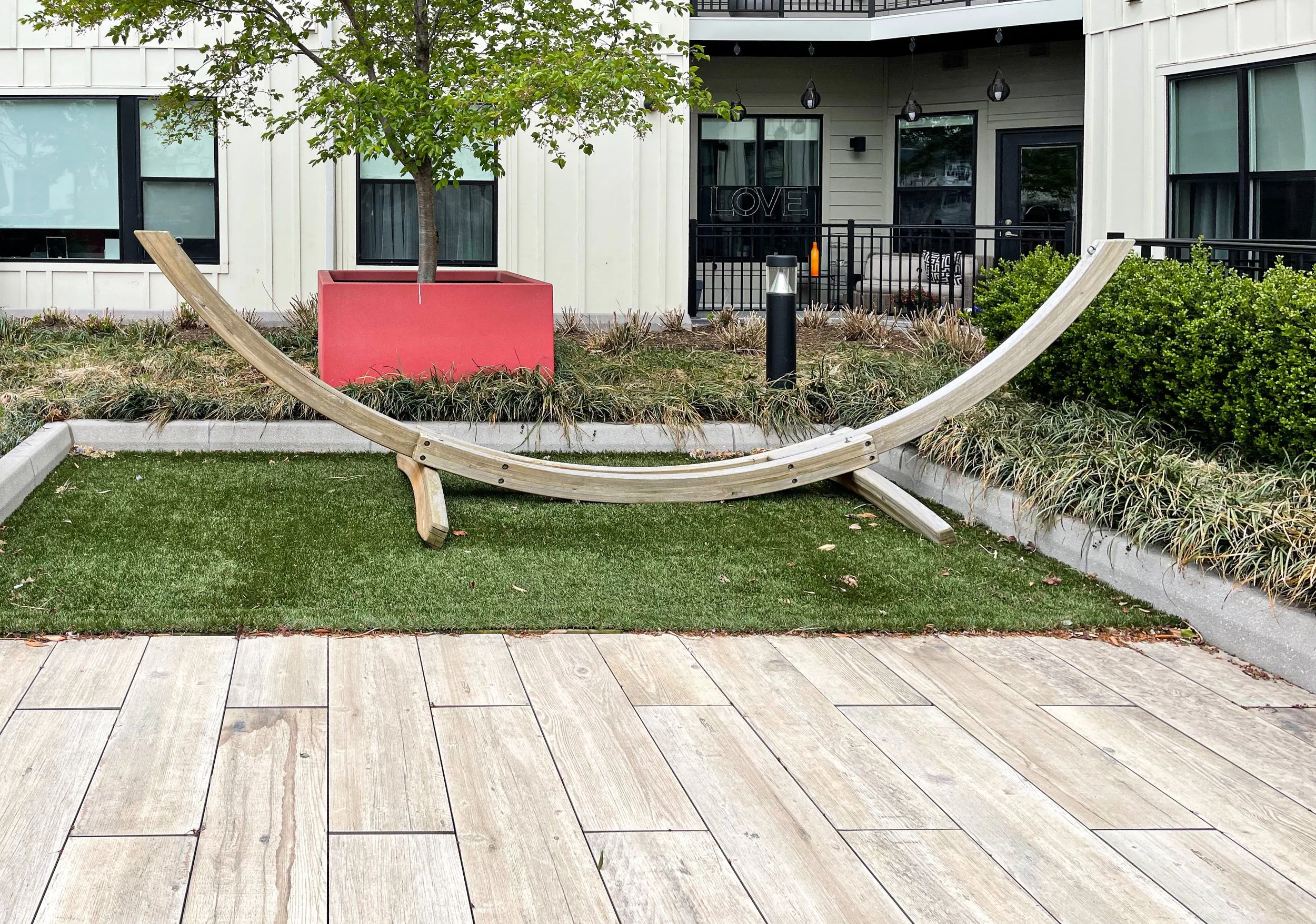 Outdoor scene with a curved wooden bench on a patch of artificial grass, surrounded by plants, with a building and balcony in the background. The building has large windows, and there's a potted tree in a red planter.