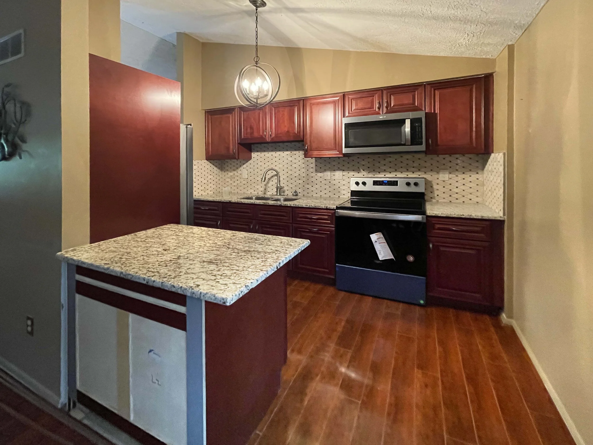 Kitchen with dark wood cabinets, granite countertops, stainless steel microwave and drawer oven, small granite island, beige walls, and hardwood floors.
