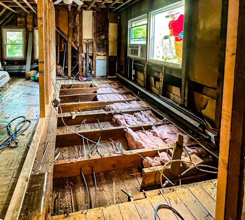 Interior of a house under renovation, with exposed floor joists, insulation, and electrical wiring visible. A window shows a person outside wearing a red shirt and yellow pants.