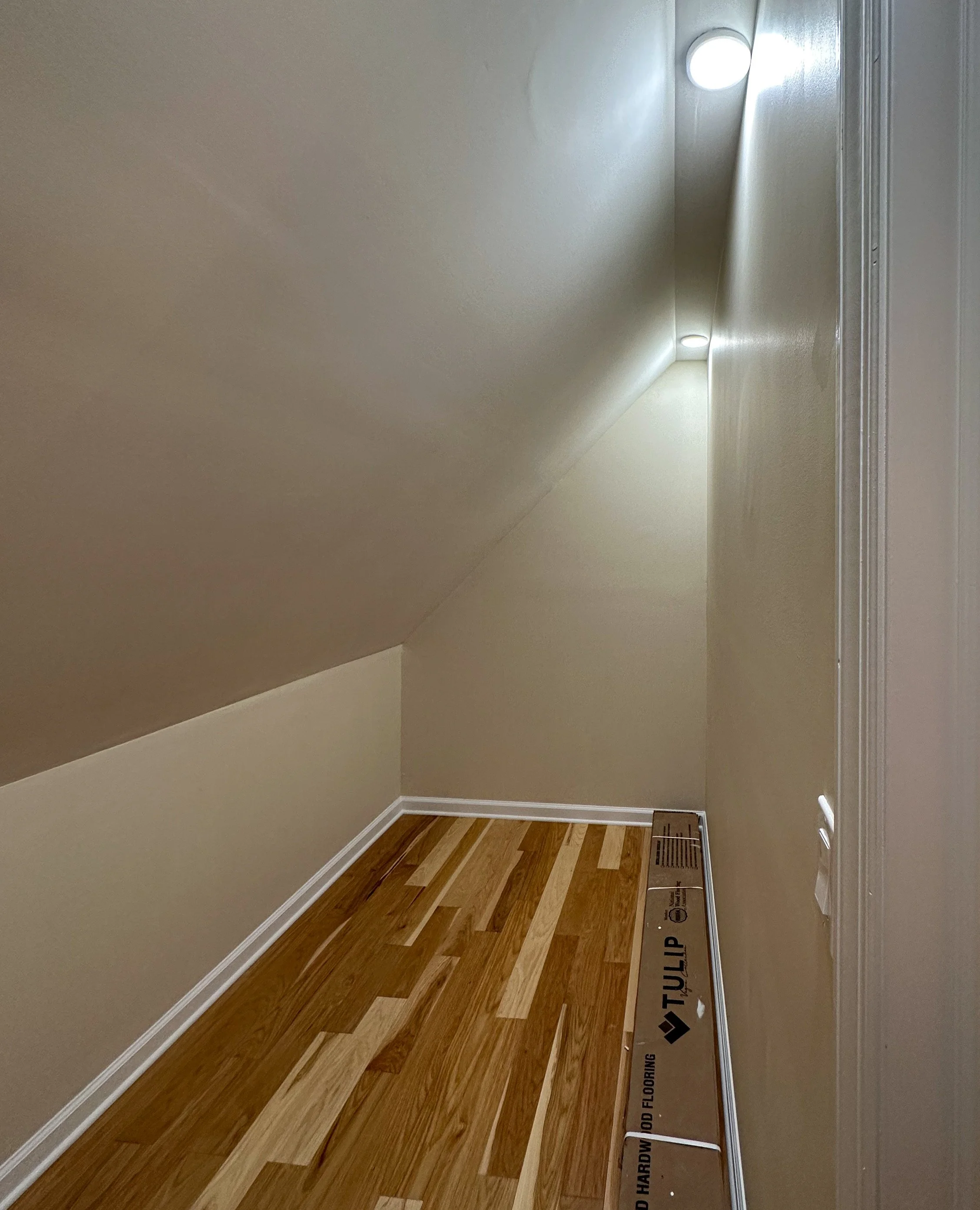 Empty attic space with sloped ceiling, hardwood flooring, beige walls, two ceiling lights, and a floor register.