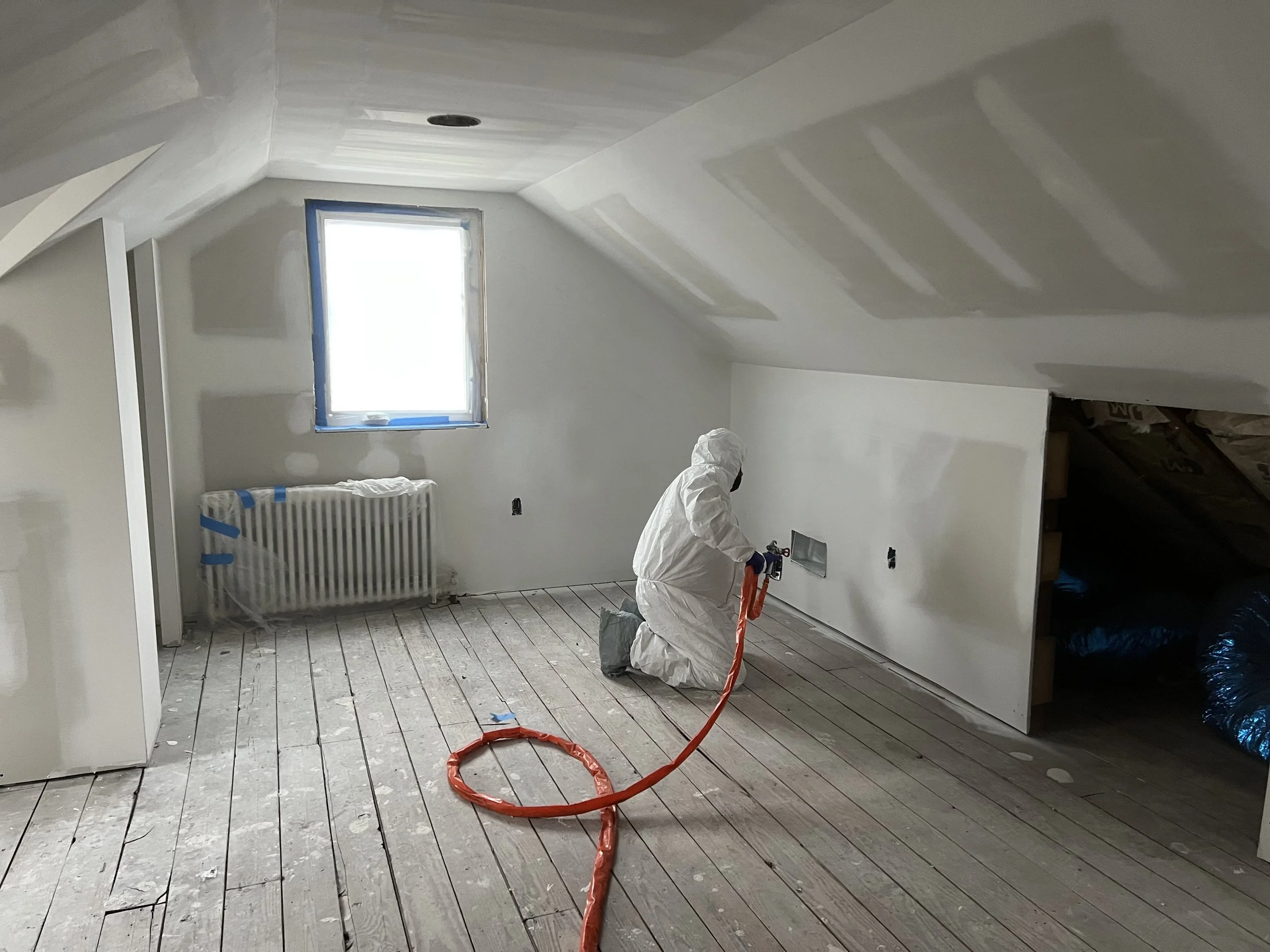 A person in protective white clothing kneeling and using a spray tool in a room under construction with white walls, a window, and unfinished wood flooring.