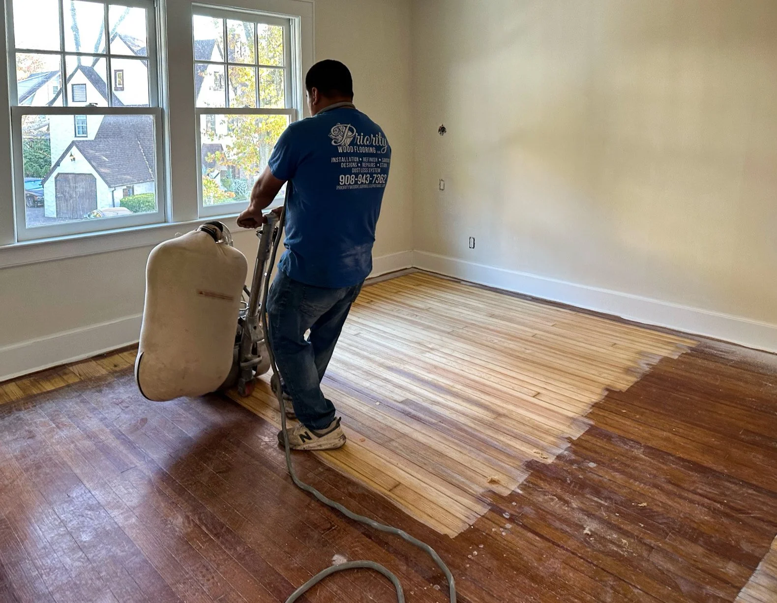 A person is sanding or polishing a wooden floor in a room with large windows, partially revealing a light hardwood floor beneath a darker finish.
