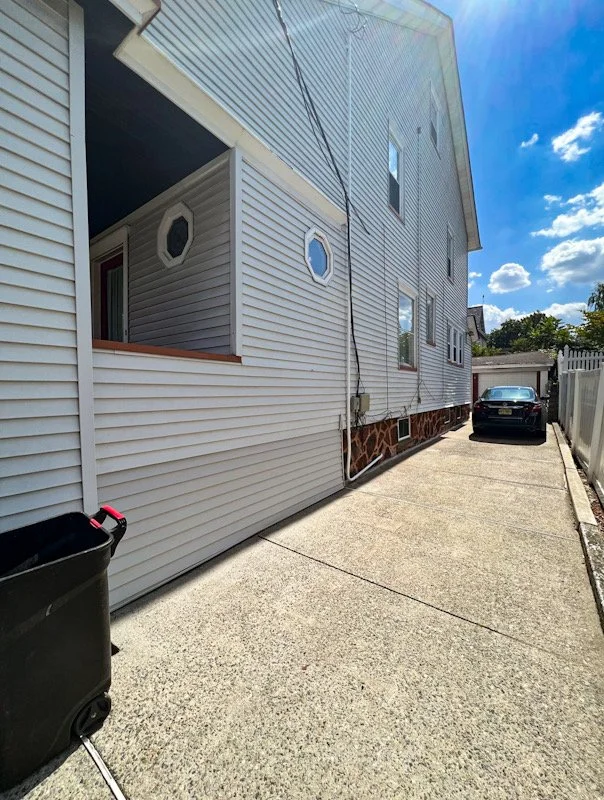 Side view of a two-story house with white vinyl siding, octagonal windows, and a stone foundation. There is a driveway with a black car parked at the end and a black trash bin on the left. A clear blue sky with some clouds is overhead.