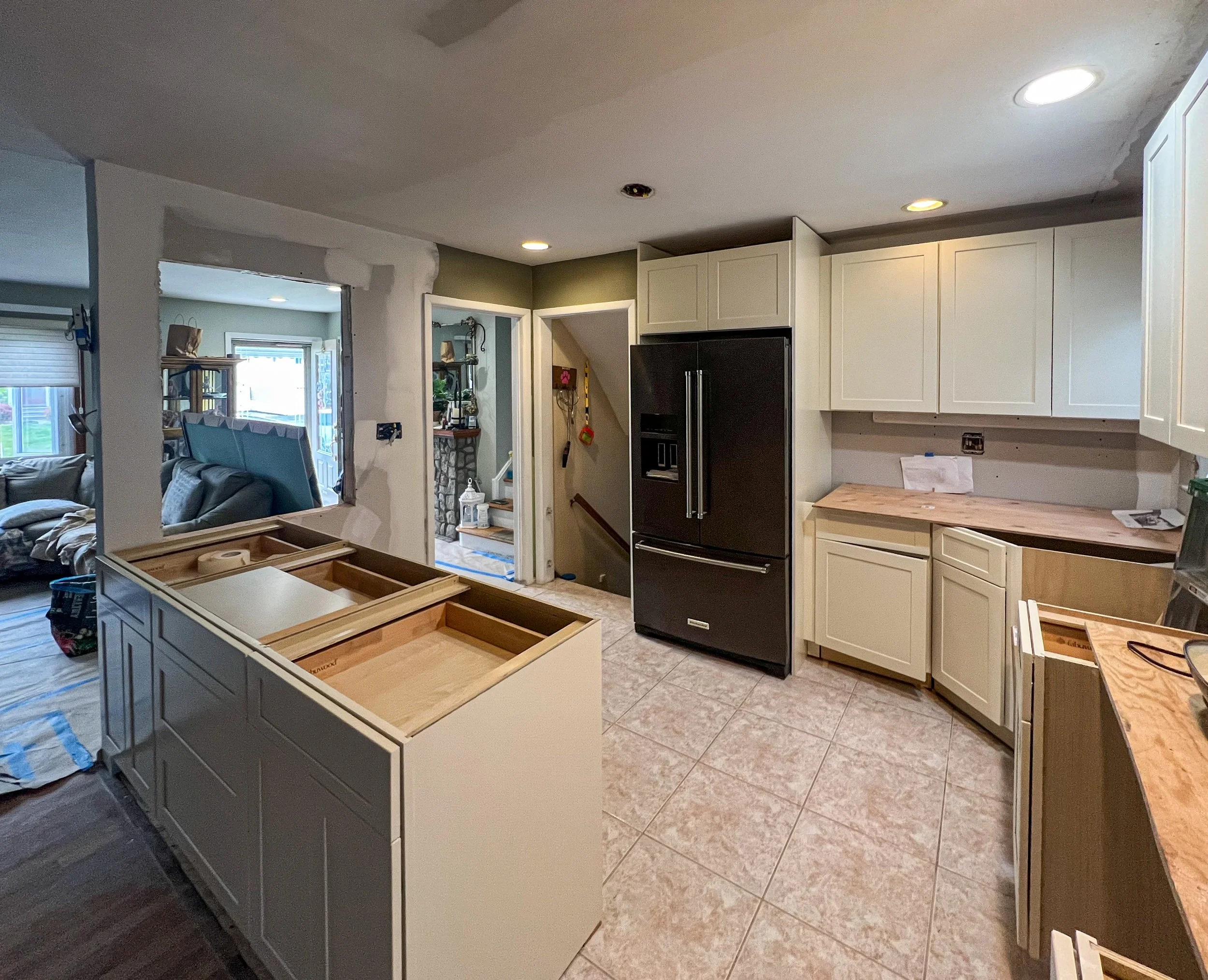 Kitchen under renovation with white cabinets, a black refrigerator, and unfinished countertops. An island with an open top is being assembled in the foreground, and the adjacent living room is visible through a partial wall opening.