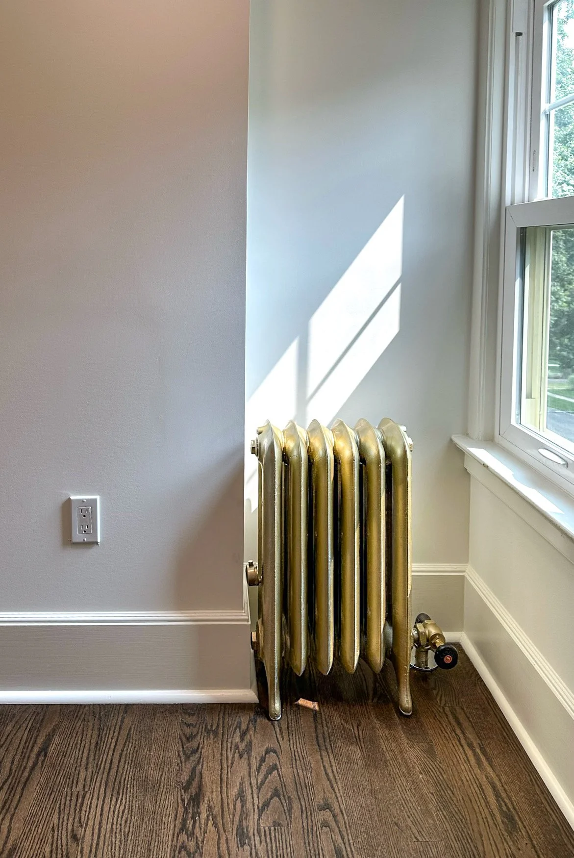 A brass radiator next to a window with sunlight casting a shadow on the wall, located in a room with a brown wooden floor and a gray wall with an electrical outlet.