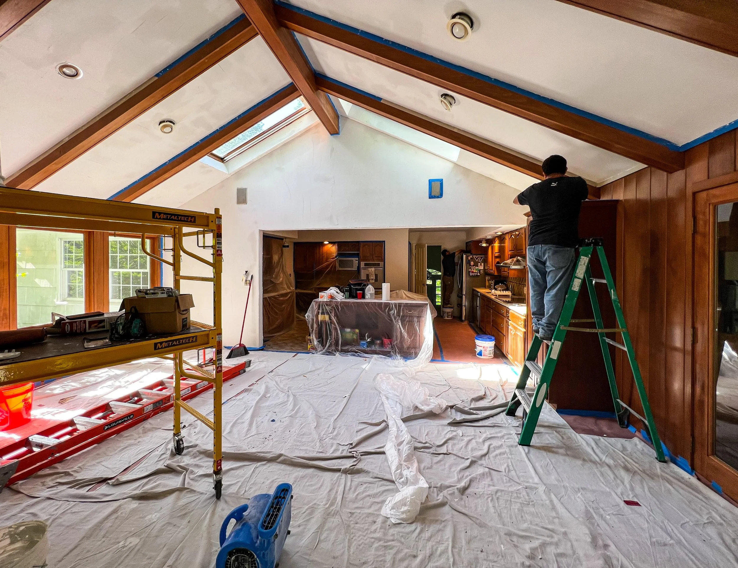 People working on home renovation inside a room with sloped ceiling, with tools and plastic coverings, one person standing on a ladder and another in the background.