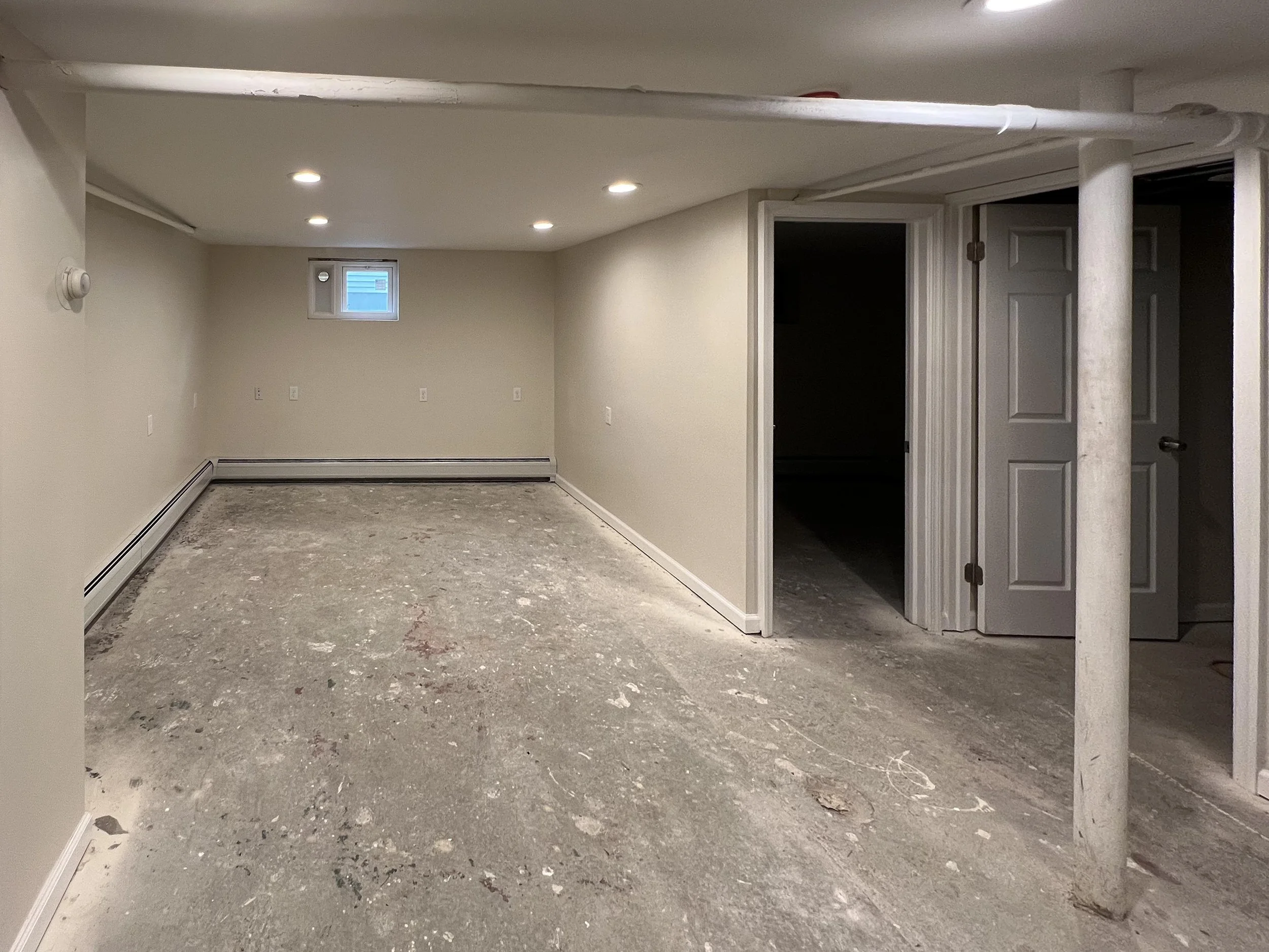 Basement under renovation with unfinished floor, beige walls, small window, and several white doors, some open, revealing storage or utility rooms.