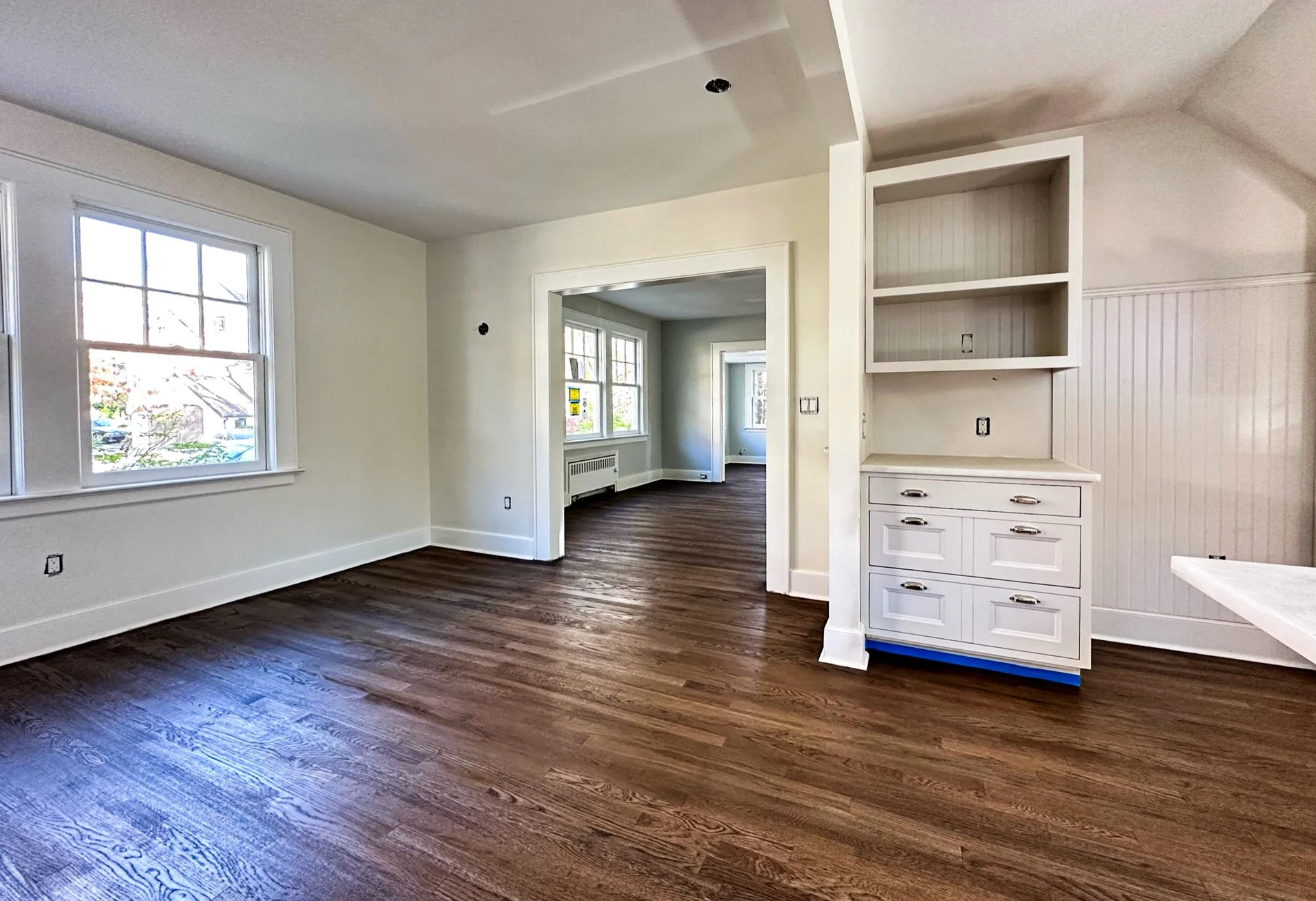 Empty room with hardwood floors, white walls, large windows, and an open kitchen area with white cabinetry.