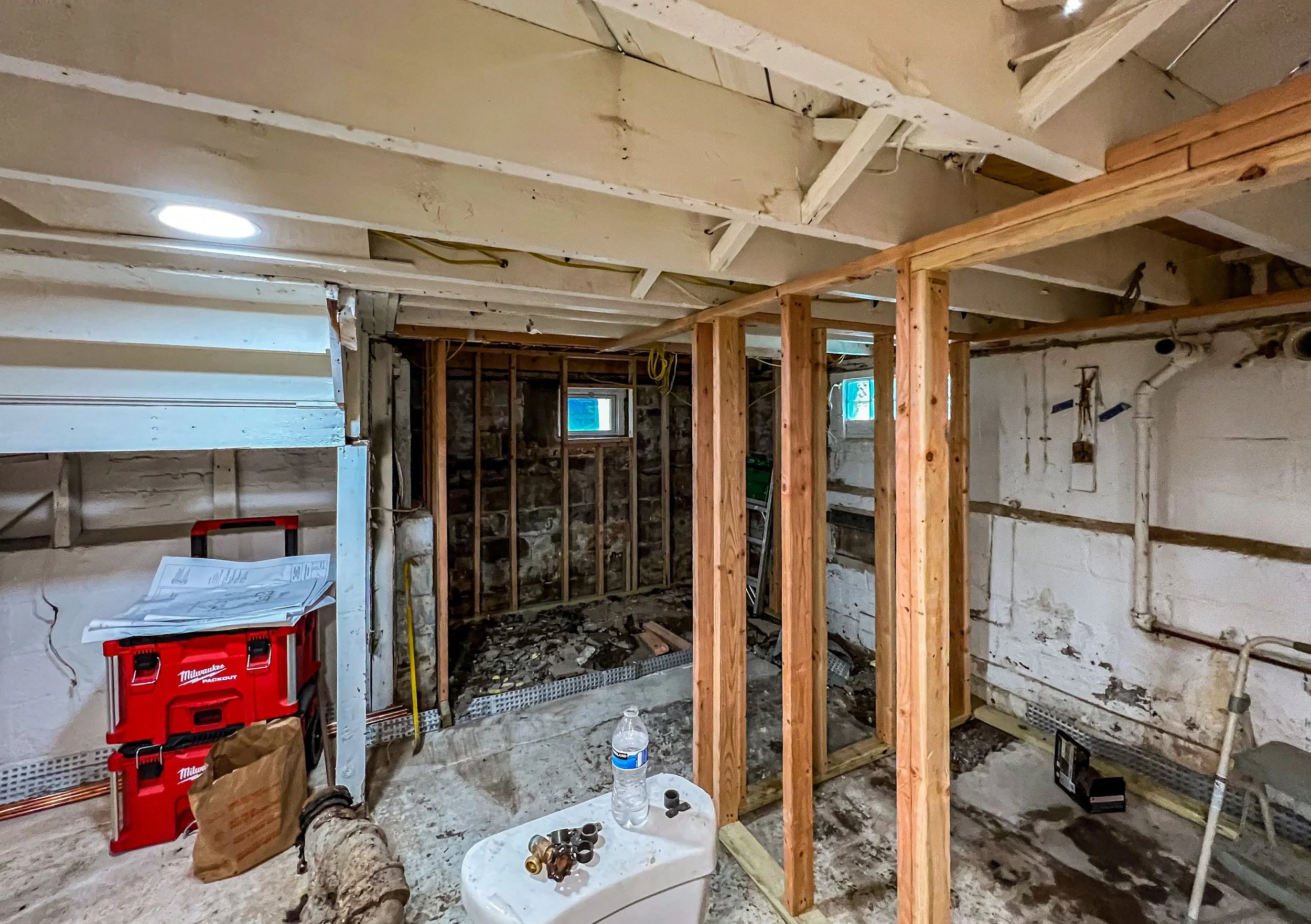 Bathroom under renovation with exposed wooden framing, construction tools, and debris, including a water bottle on a small table.