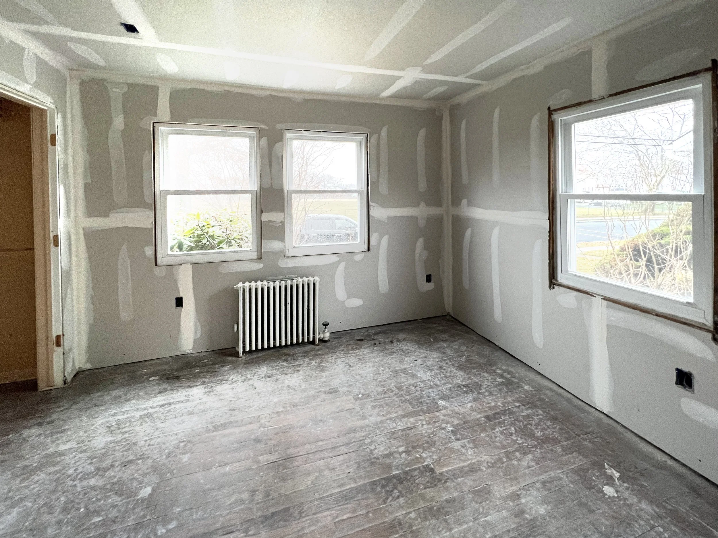 Interior of a room under construction with drywall partially finished, three windows, an old radiator under the middle window, exposed floor, and unpainted wall edges.