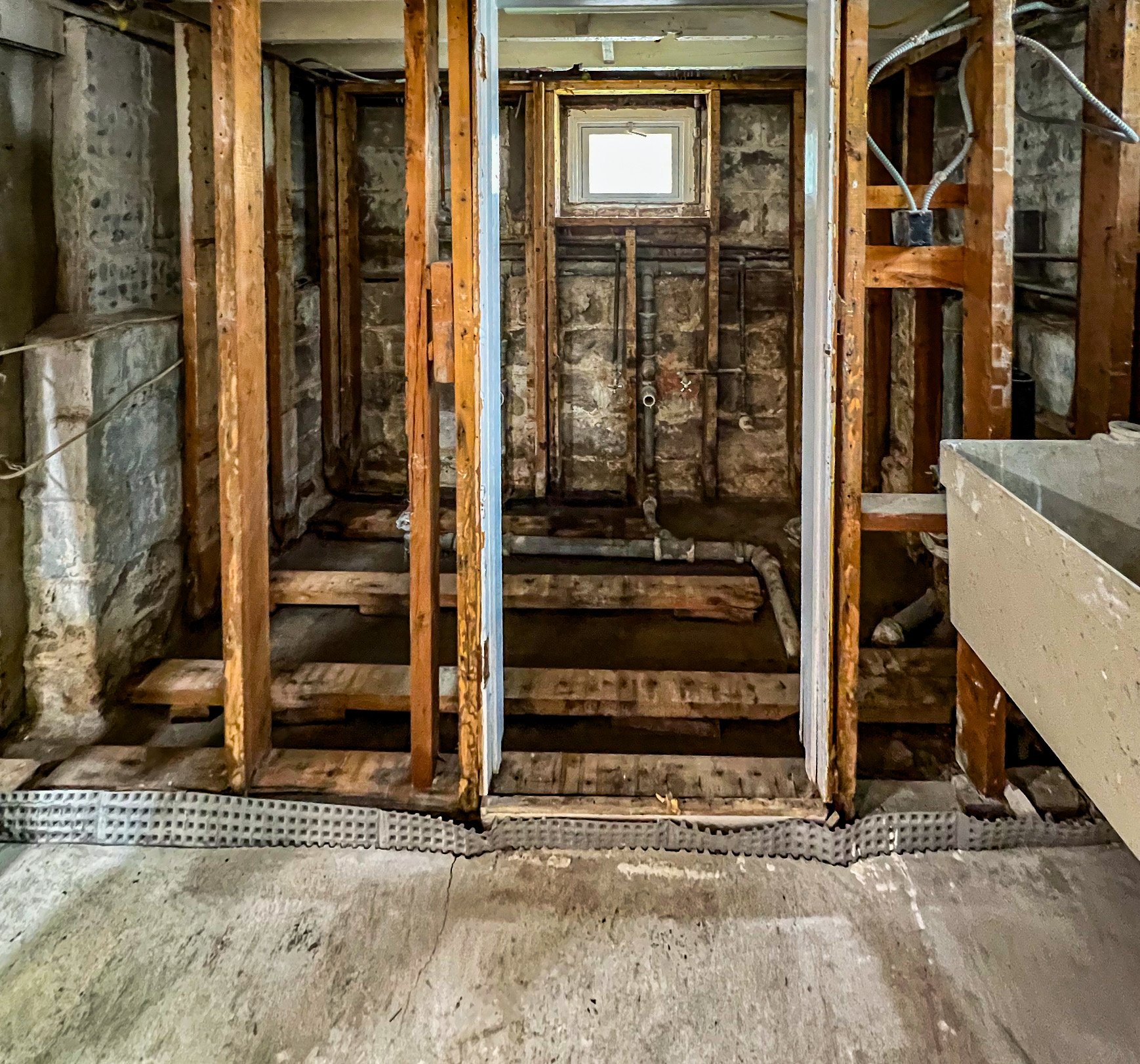 The photo shows a bathroom under renovation with exposed wooden studs, a small window, and plumbing pipes. The floor is unfinished, and construction materials are present.