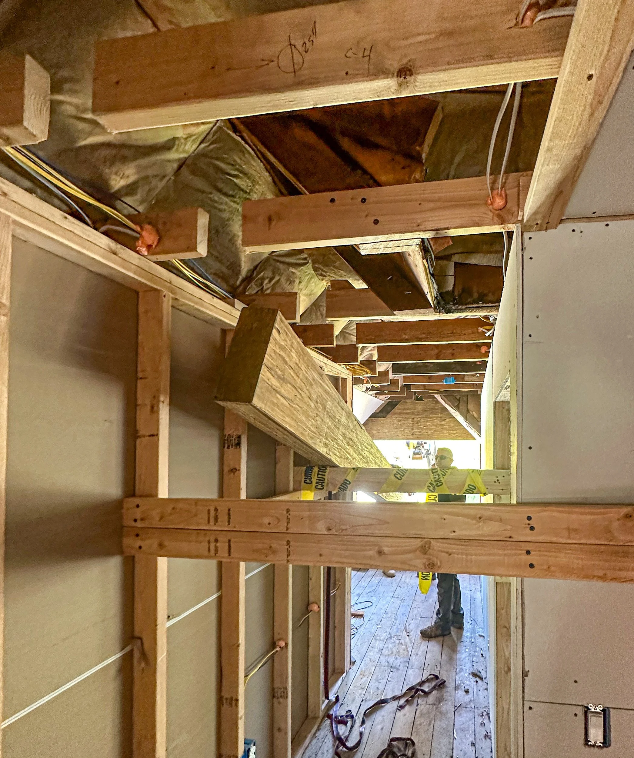Interior view of a construction site showing wooden framing and insulation, with workers in the background.