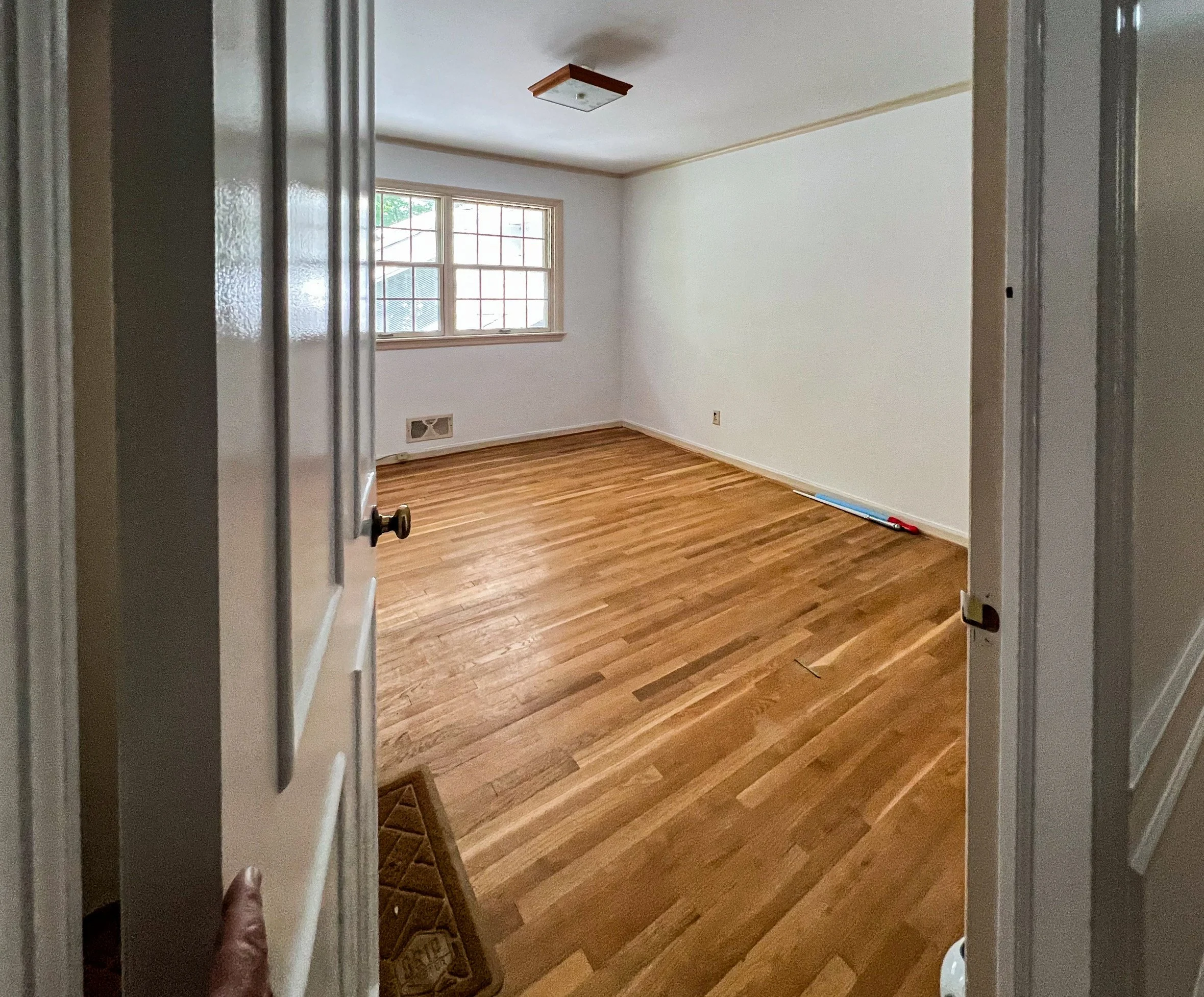 Empty room with wooden floors, white walls, a large window, and a ceiling light fixture. Part of the door is visible from which the photo was taken.