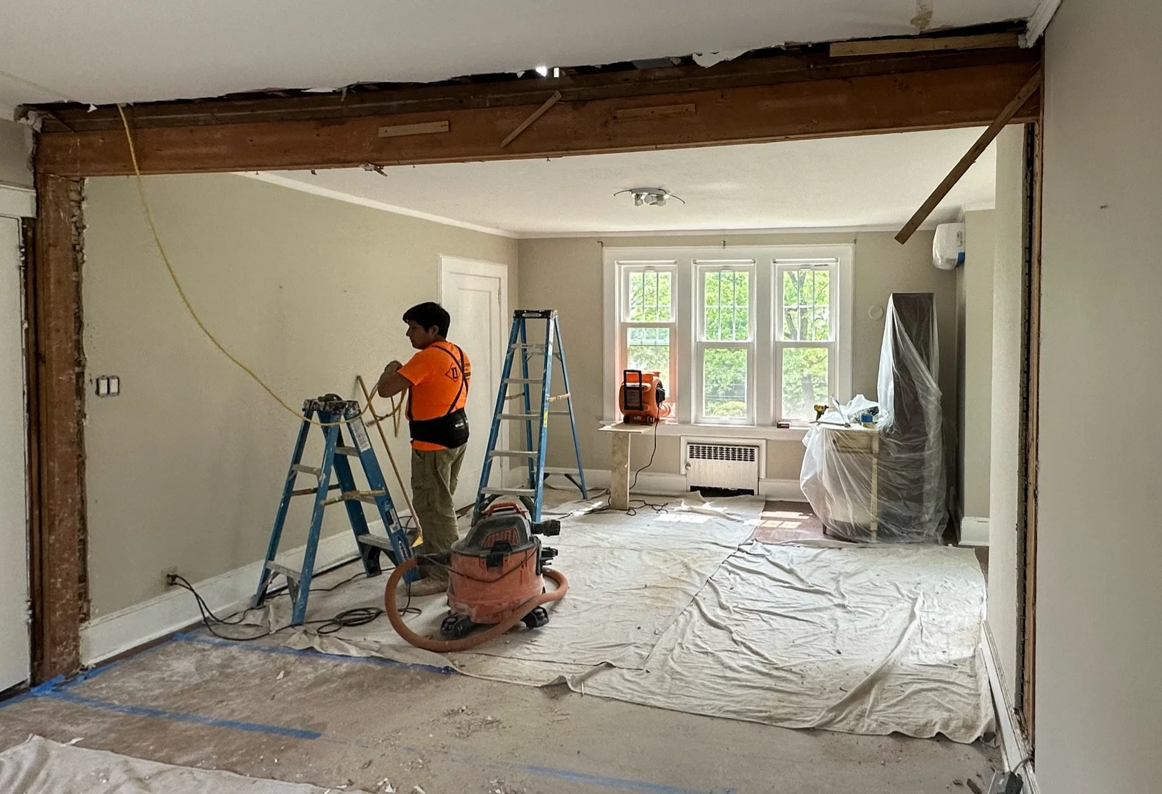 A construction worker in an orange shirt is working on home renovation inside a room with large windows, ladders, and construction tools, with plastic covering furniture and a floor sheet on the ground.