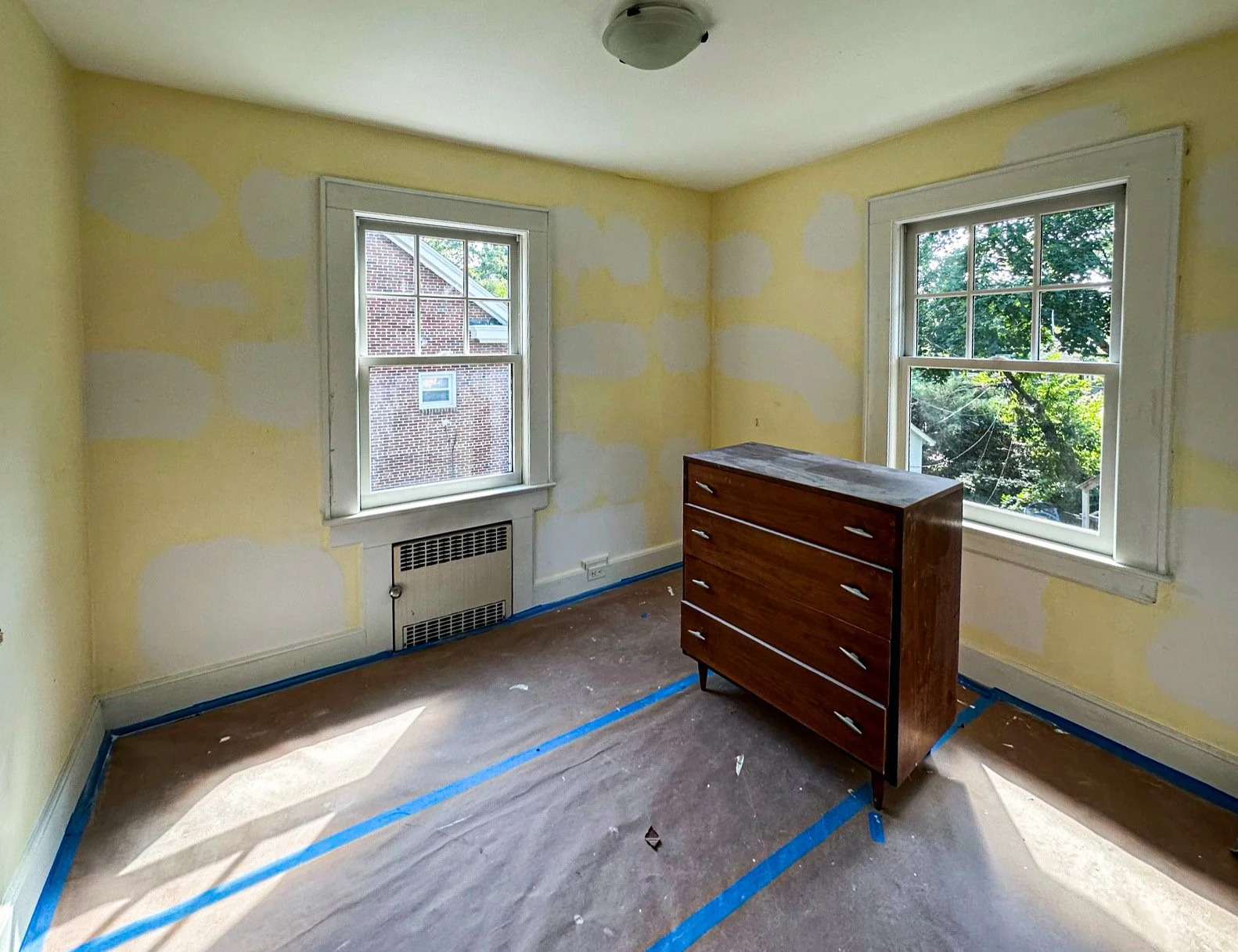 Empty room with yellow walls and white trim, two windows showing trees and a brick building outside, a wooden dresser, and painter's tape on the floor.