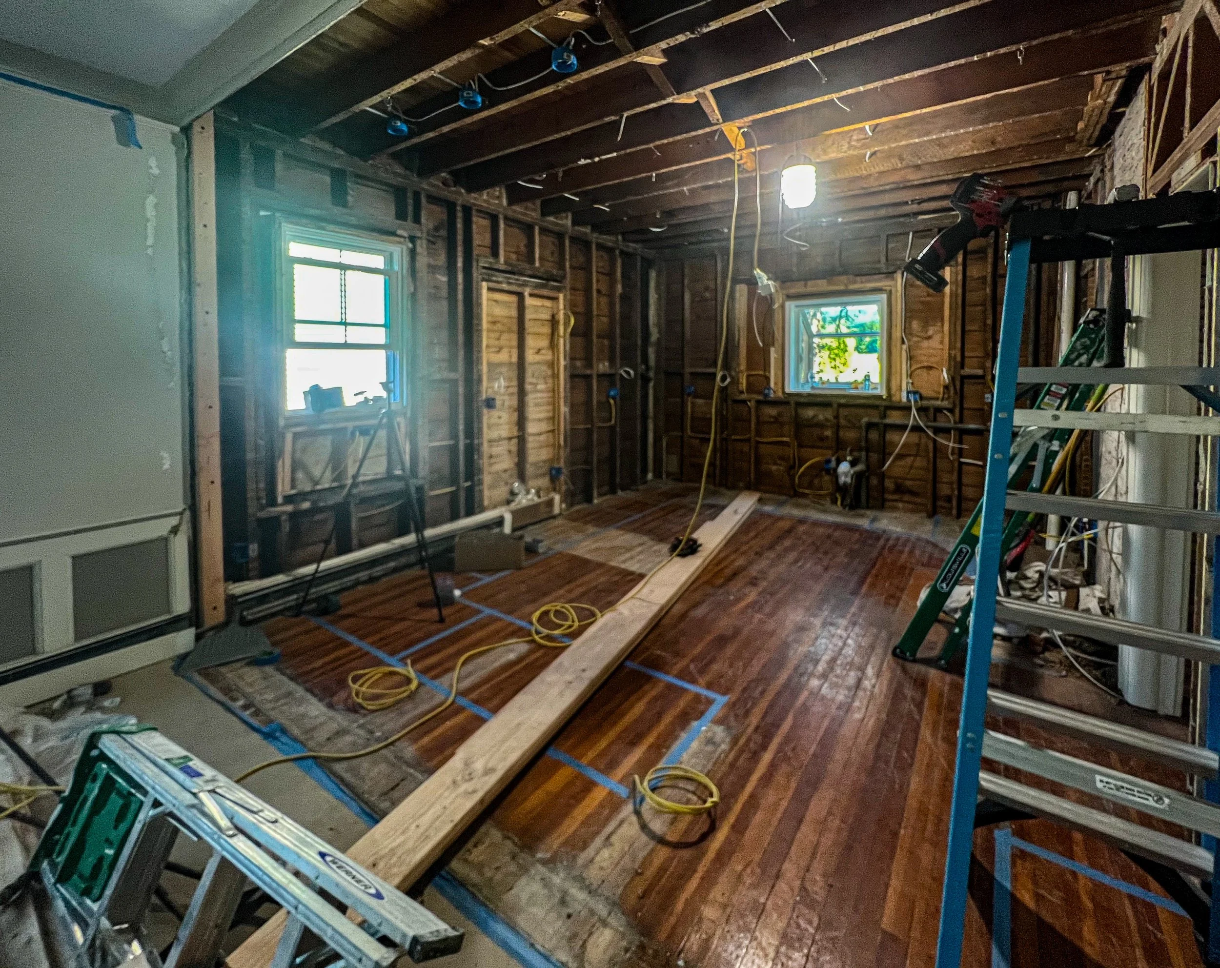 Room under renovation with exposed wooden walls and ceiling beams, partially finished hardwood flooring, work tools such as a ladder, extension cords, and a plank, two windows letting in natural light, some electrical wiring, and construction materia