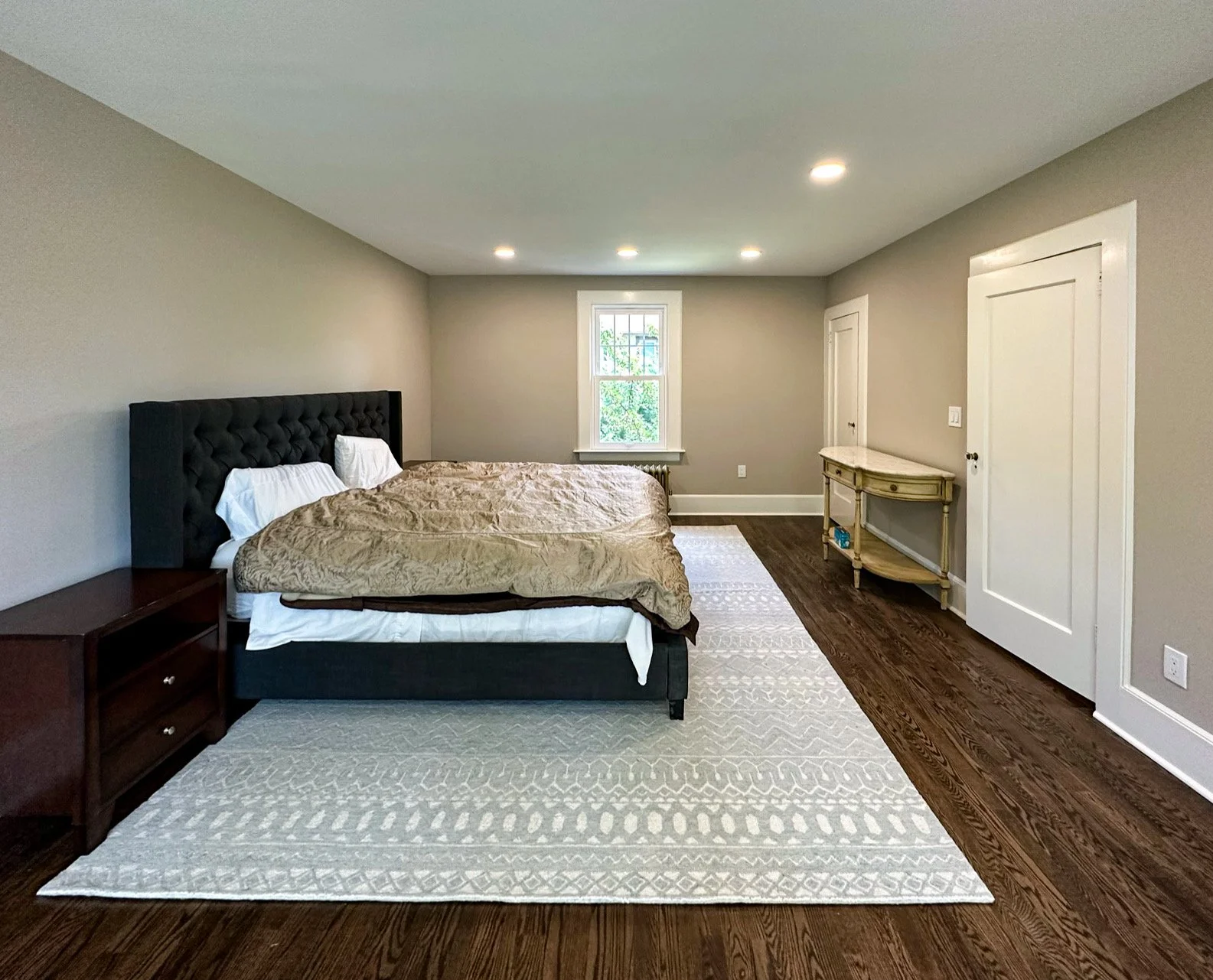 A spacious bedroom with a large bed, a dark tufted headboard, white pillows, and beige bedding. There's a nightstand on the left, a white patterned rug, and a small vintage-style console table on the right wall. The room has gray walls, wood flooring