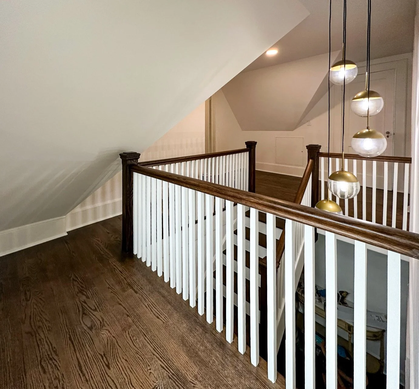 Interior view of a staircase landing with wooden flooring, white railing with dark wood handrail, and modern hanging light fixtures with white and gold accents.