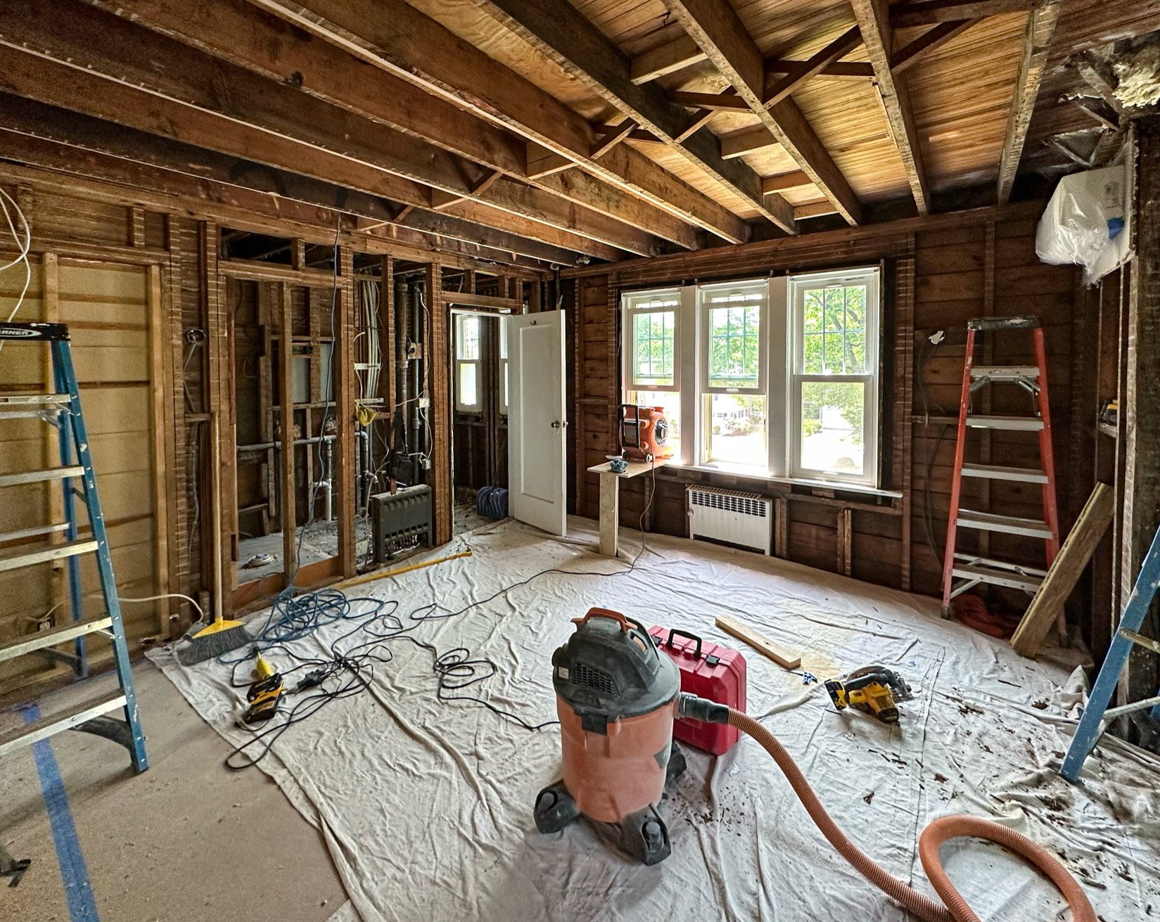 Interior of a room under renovation with exposed wooden studs and ceiling beams, construction tools on a tarp-covered floor, and a window showing trees outside.