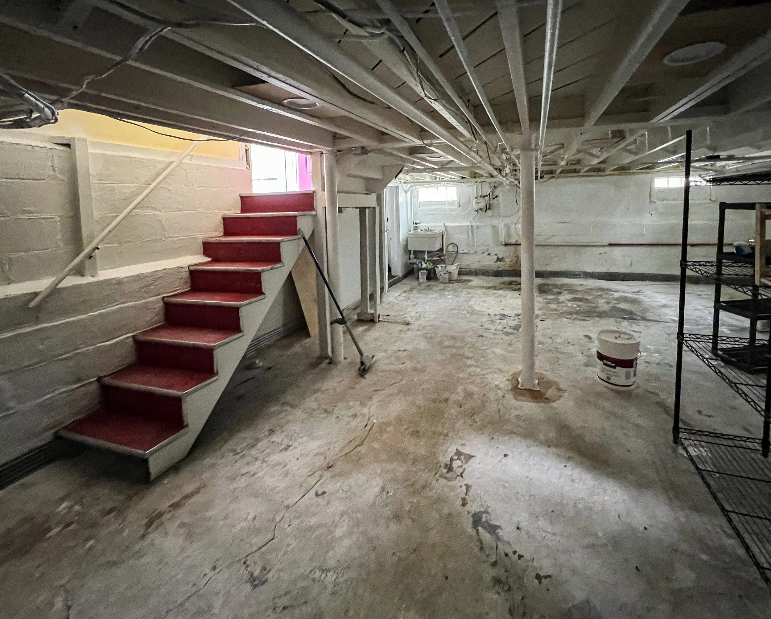Basement with unfinished concrete flooring, exposed ceiling pipes, a small staircase with red steps, a window letting in natural light, a laundry area with a washing machine, some buckets and a paint bucket, and metal shelving on the right.