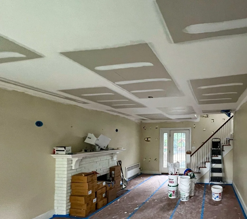 Living room under renovation with drywall ceiling, fireplace, and staircase, construction materials, and tools scattered around.