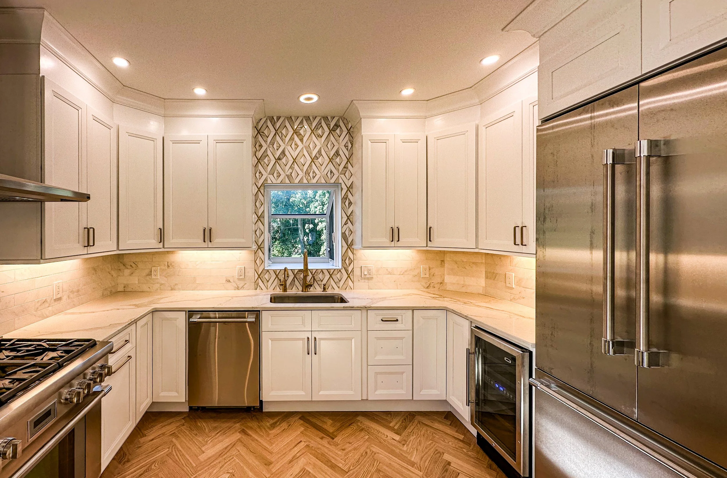 Modern kitchen with white cabinets, marble countertops, gold hardware, stainless steel appliances, a window with a geometric tile backsplash, and hardwood floors.