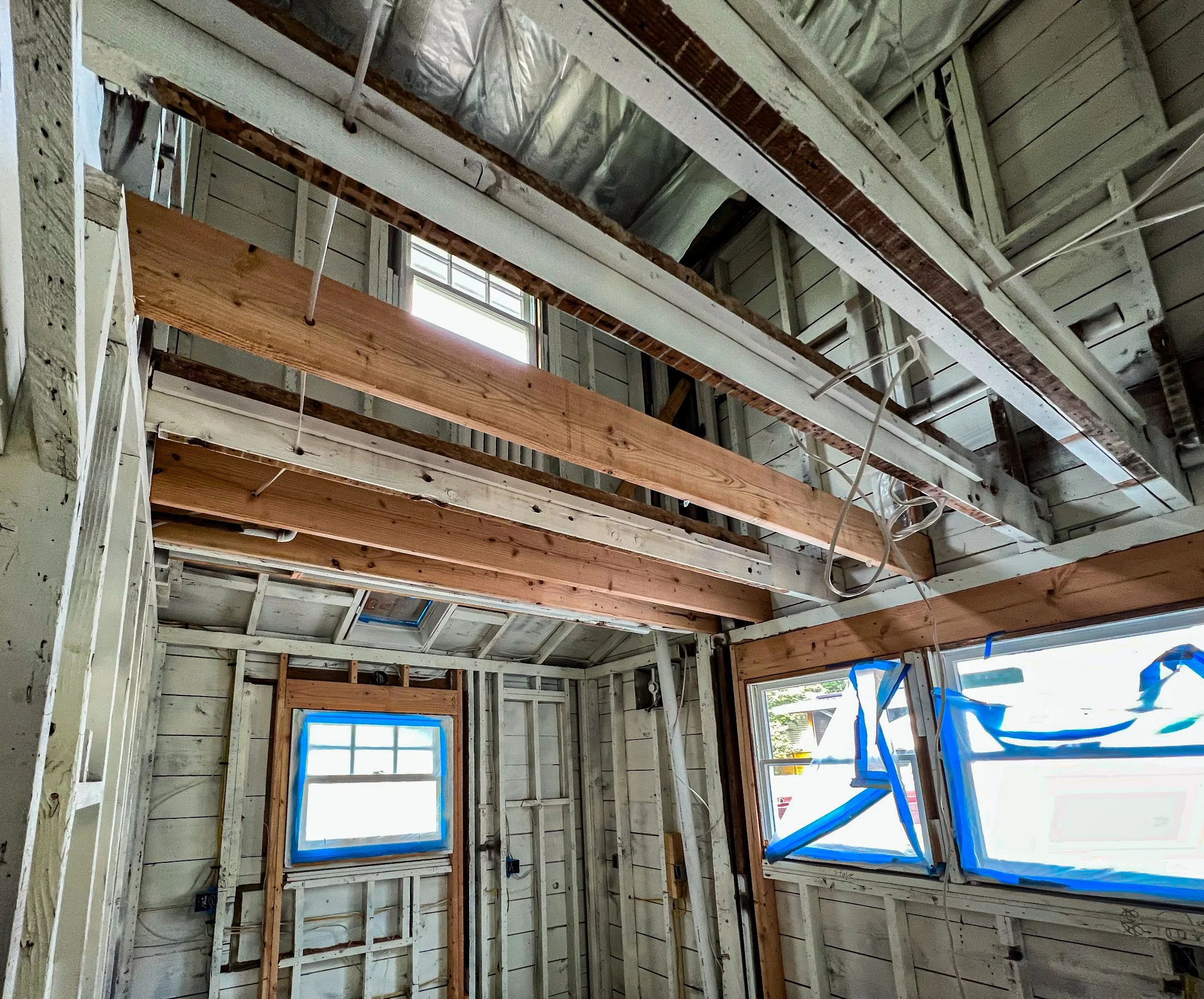 Interior of a house under construction with exposed framing, insulation, and wiring, including windows covered with blue painter's tape.