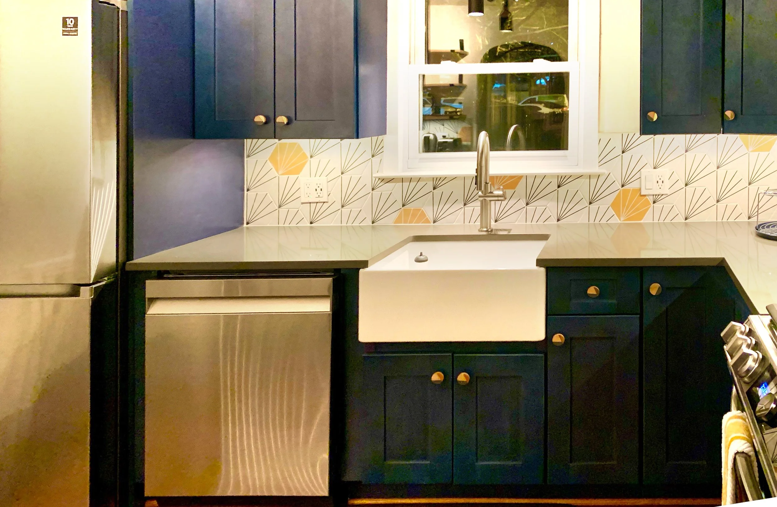 Kitchen with navy blue cabinets, white farmhouse sink, stainless steel appliances, geometric patterned tile backsplash in tan, white, and black, and a window above the sink.