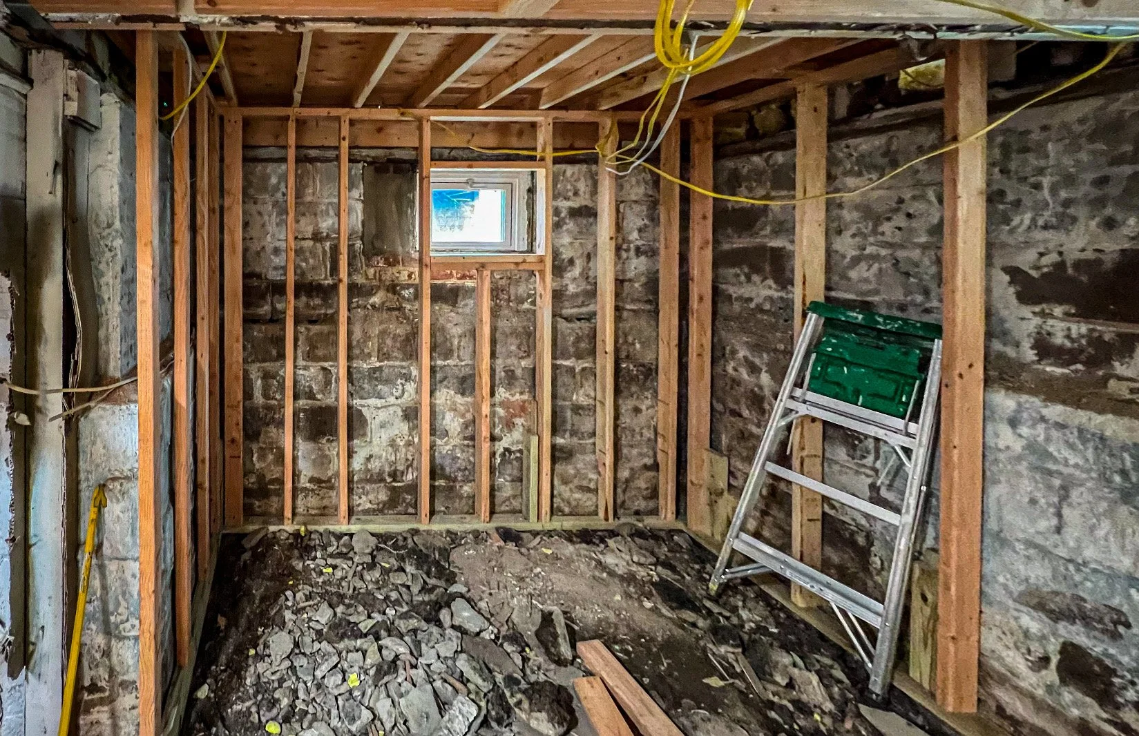 Construction room with wooden framing, exposed stone walls, a small window, a metal stepladder, and a pile of rocks and debris on the floor.