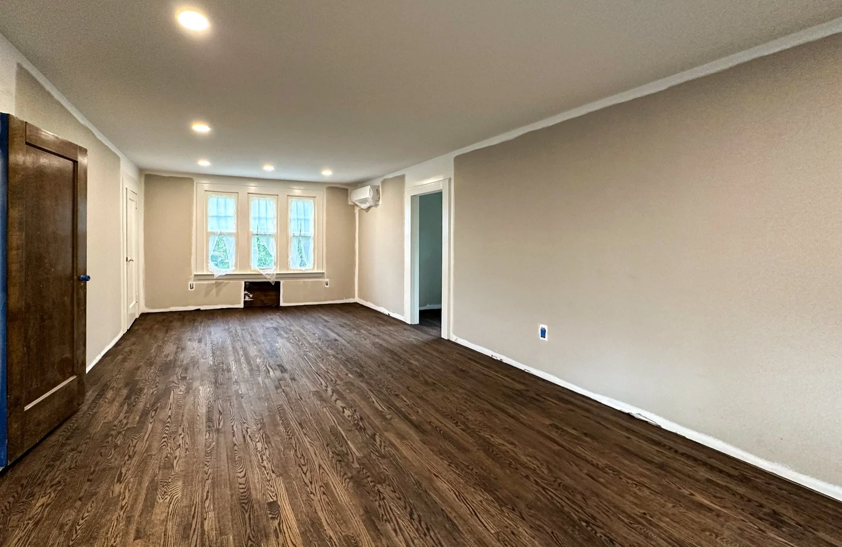 Empty living room with dark hardwood floors, beige walls, white trim, three windows, and recessed ceiling lights.