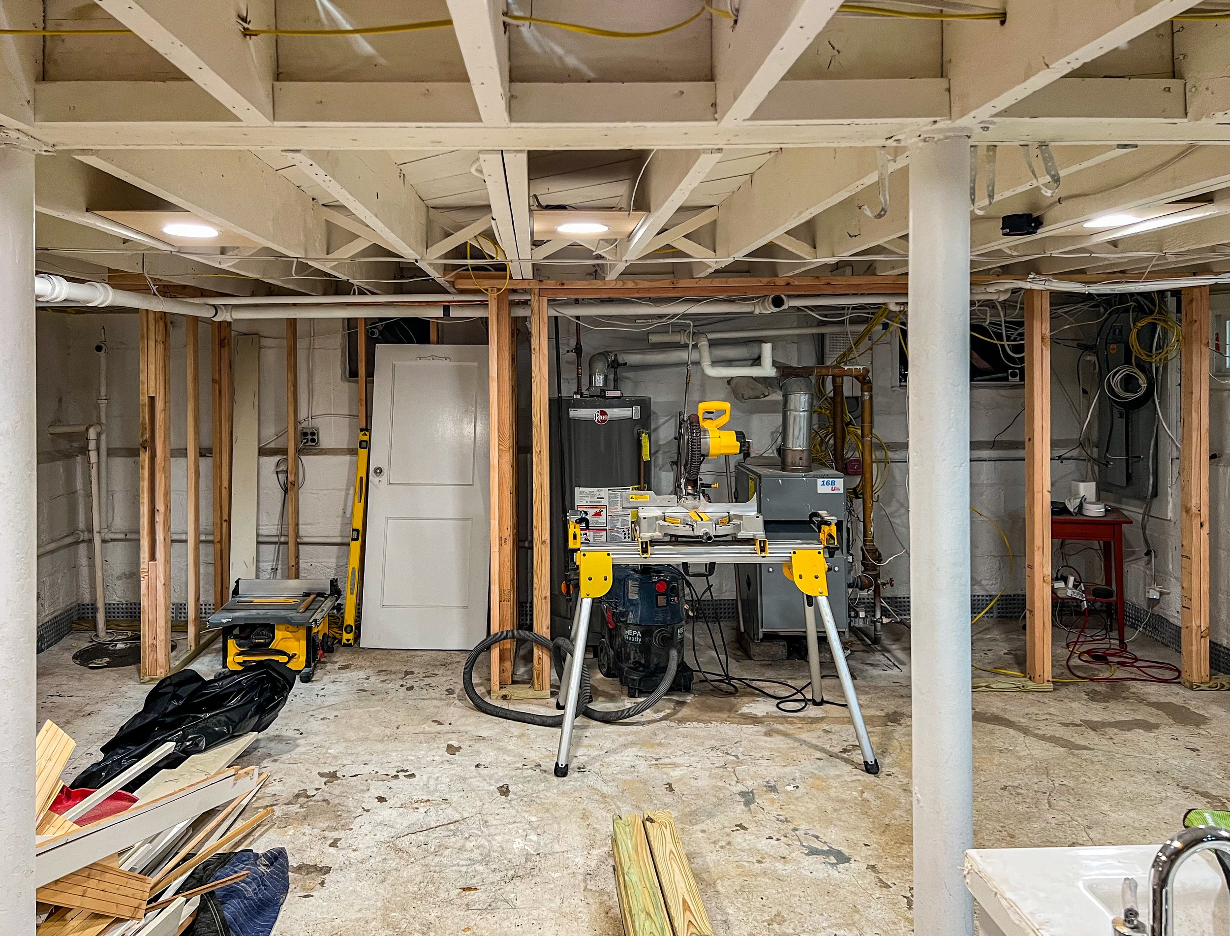 A laundry room or utility basement under construction, with exposed wooden beams, unfinished concrete floor, and various tools and equipment including a table saw, a drill, and pipes, with wiring hanging from the ceiling.