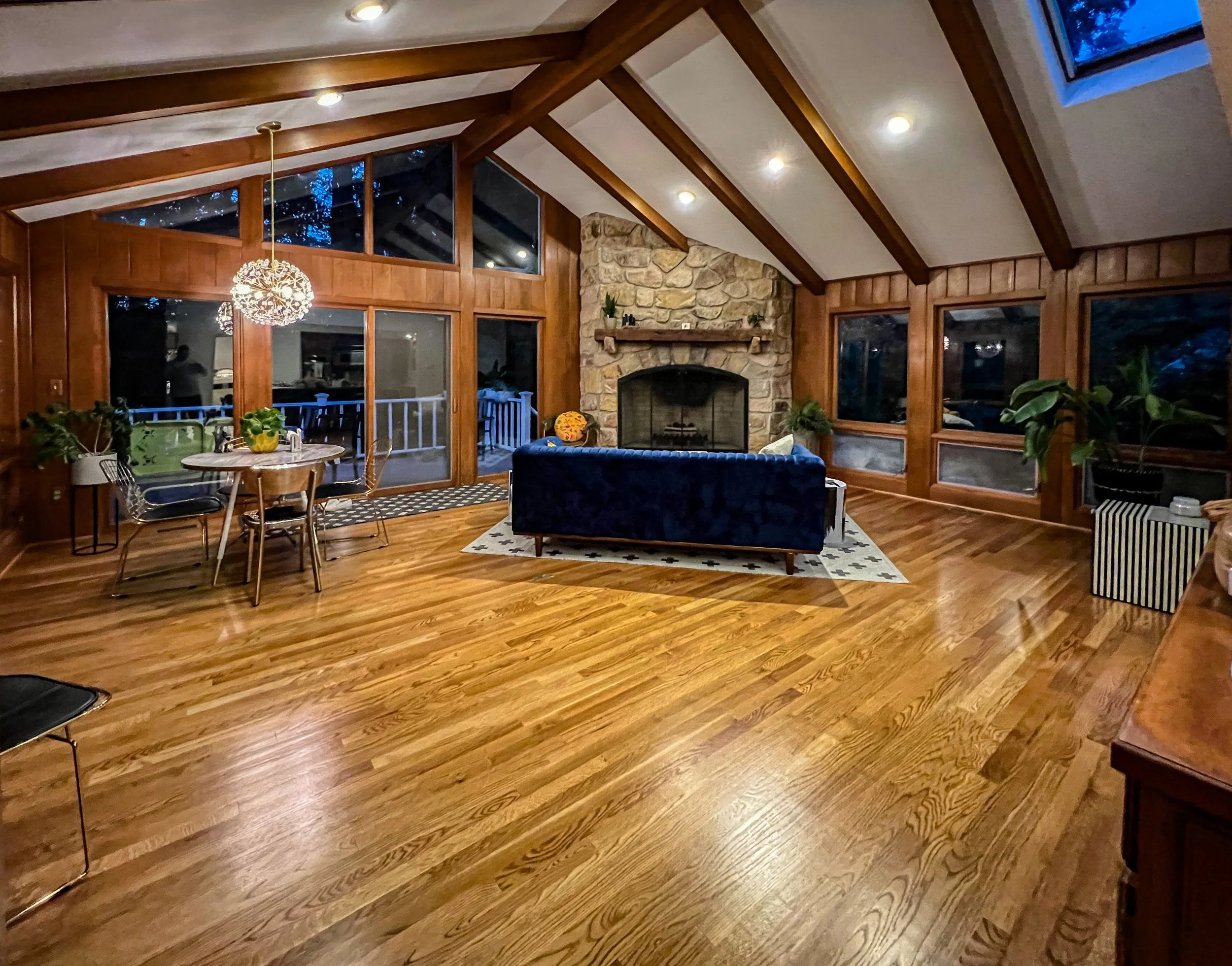 Spacious living room with hardwood floors, a stone fireplace, large windows, a round dining table with chairs, a navy velvet sofa, and indoor plants.