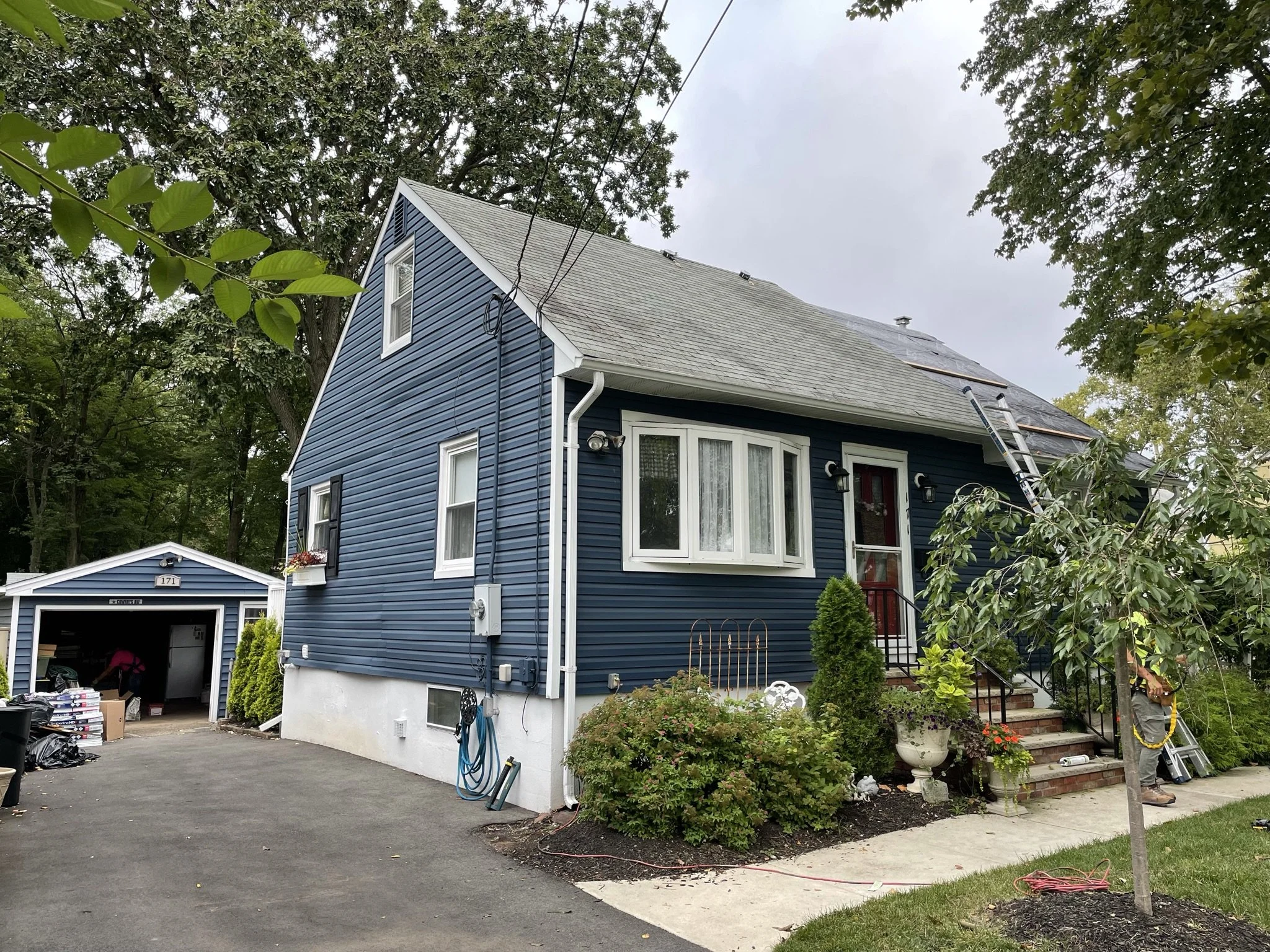 A blue house with white trim and black shutters in a residential neighborhood. A person is working near the front steps, which are decorated with plants and flowers. The house has a pitched roof, a window with a bay window feature, and a detached gar