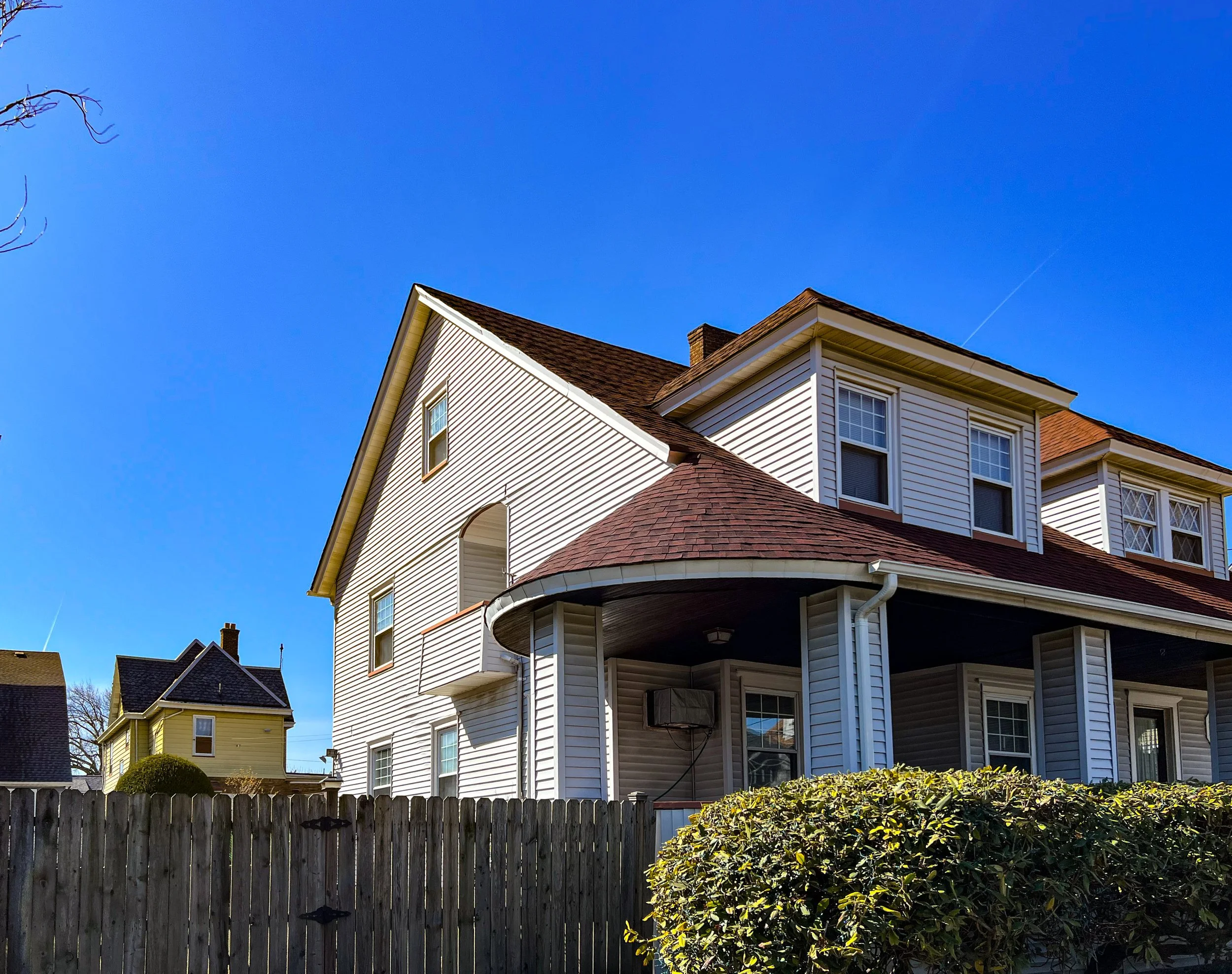 A large, white, multi-story house with brown roof shingles and a covered porch, set against a bright blue sky with a few tree branches in the upper corners.