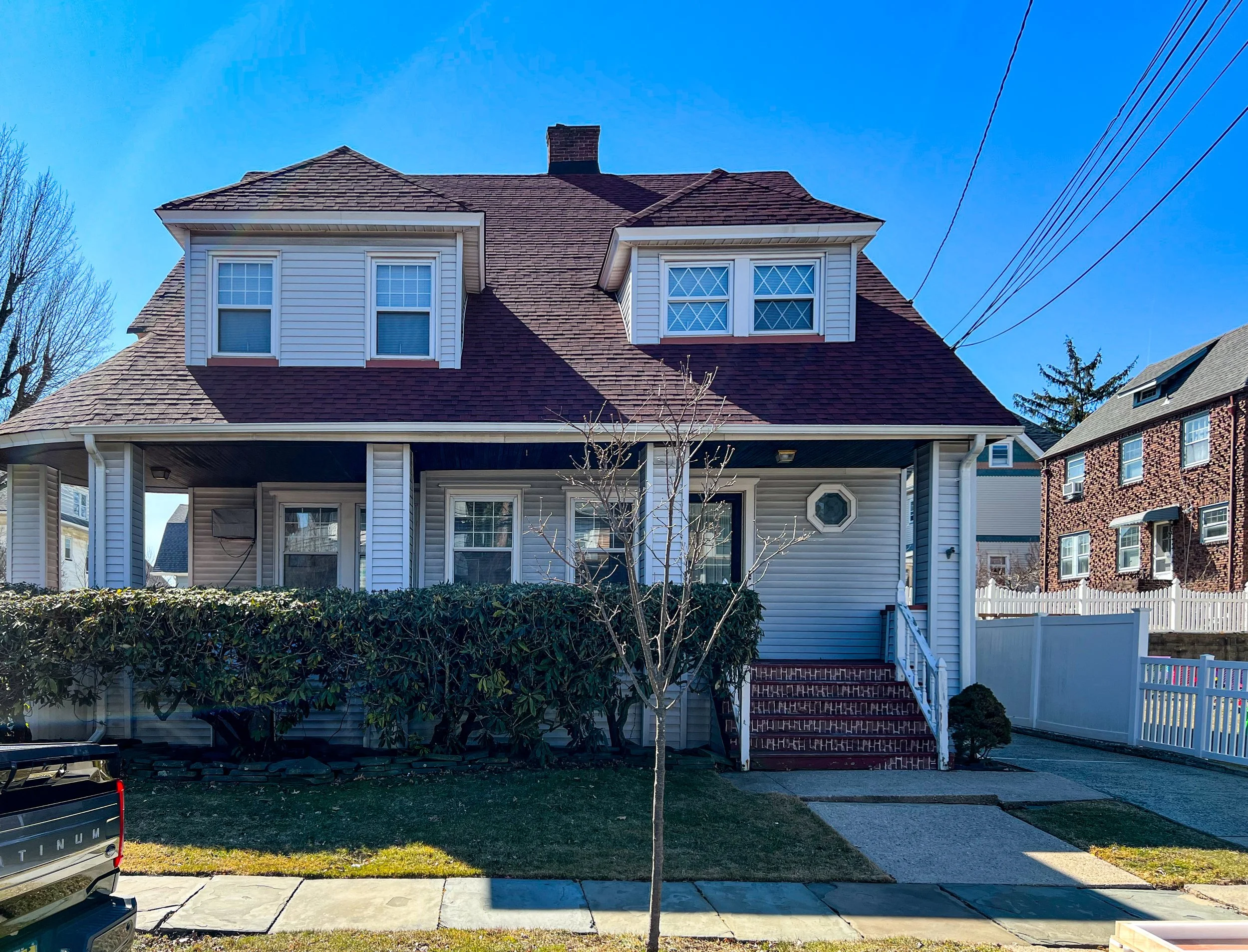 Front view of a two-story house with beige vinyl siding, a brick front porch staircase, white trim, and a brown roof, under a clear blue sky.