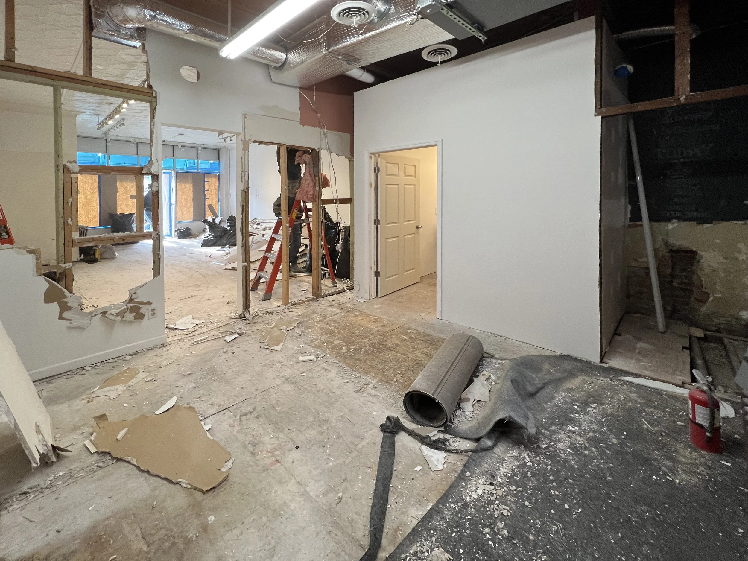 Room under renovation with construction debris, exposed framing, a worker on a ladder, and remnants of drywall and flooring.