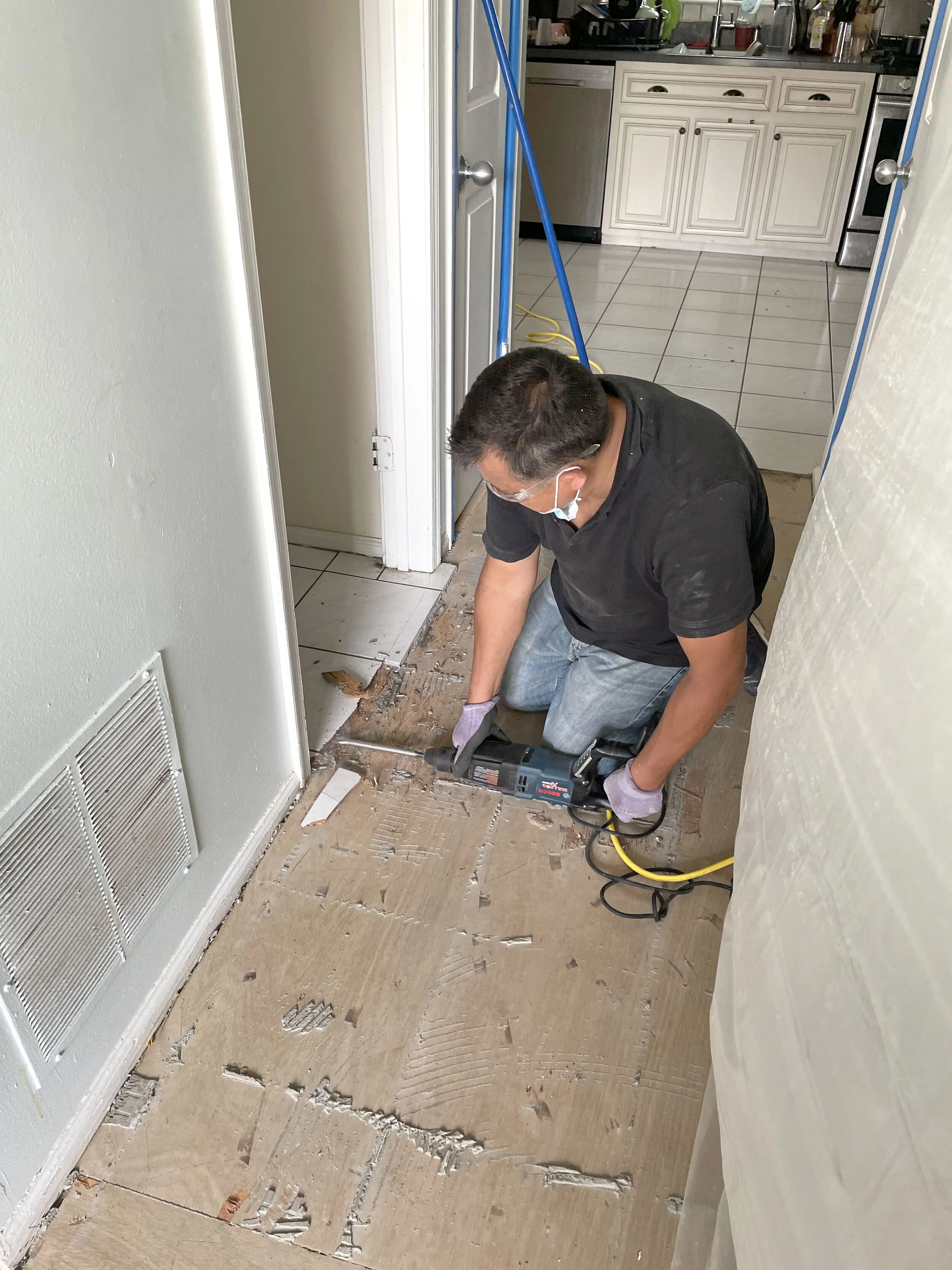 A person wearing a face mask and gloves kneels on the floor, using a power tool to remove old flooring in a kitchen area.