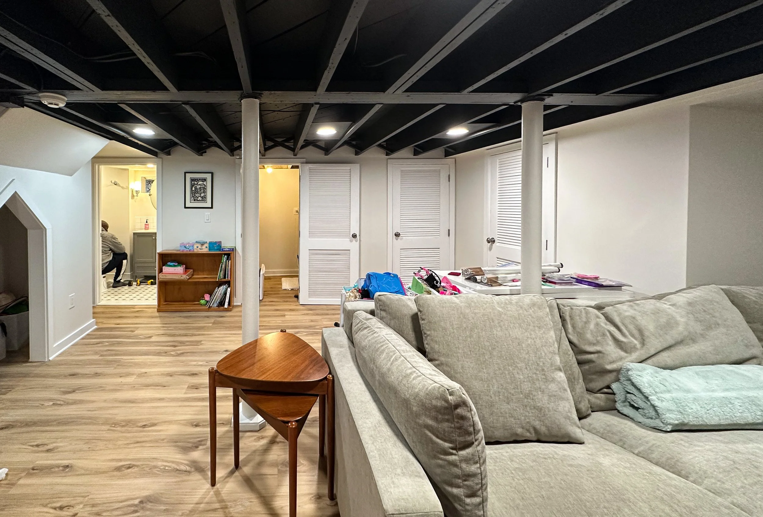 A living room with beige couch, wooden side table, and cluttered table with papers and backpacks. In the background, a person seated in a bathroom or laundry area, with doors leading to a hallway.