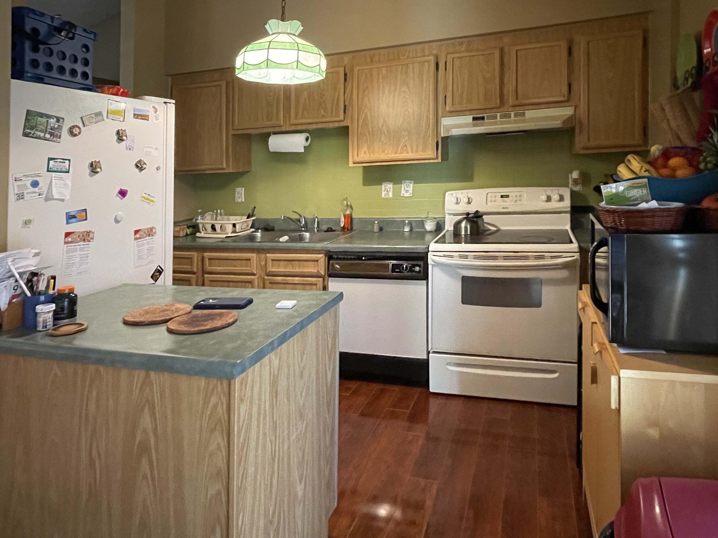 A kitchen with wooden cabinets, a white refrigerator covered with magnets and papers, a green wall, and a countertop with various items. There are three pancakes and a phone on the small island in the foreground.