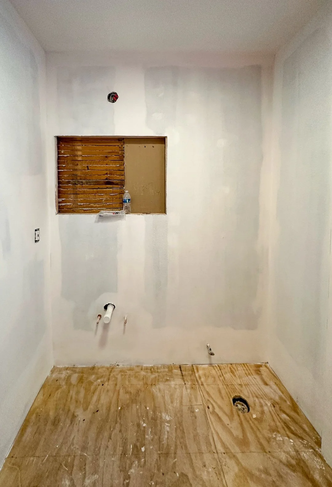 Empty room under renovation with white walls, a small window with wooden slats, exposed pipes, and a plywood floor.