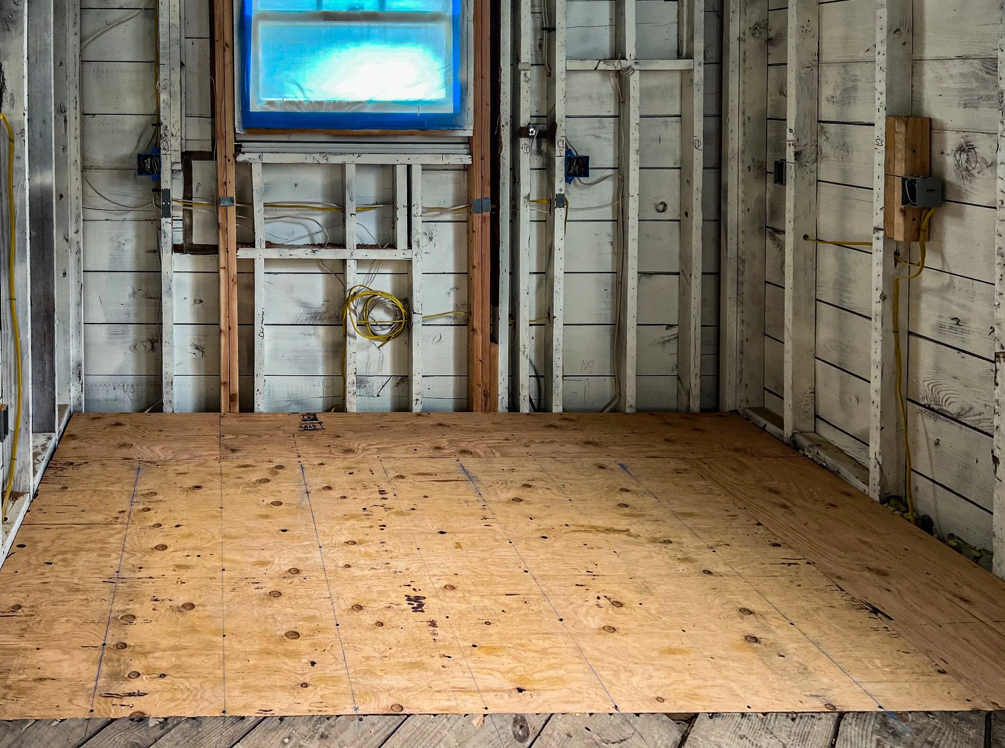 Interior of a room under construction with plywood flooring and exposed wall studs, electrical wiring, and a small window.