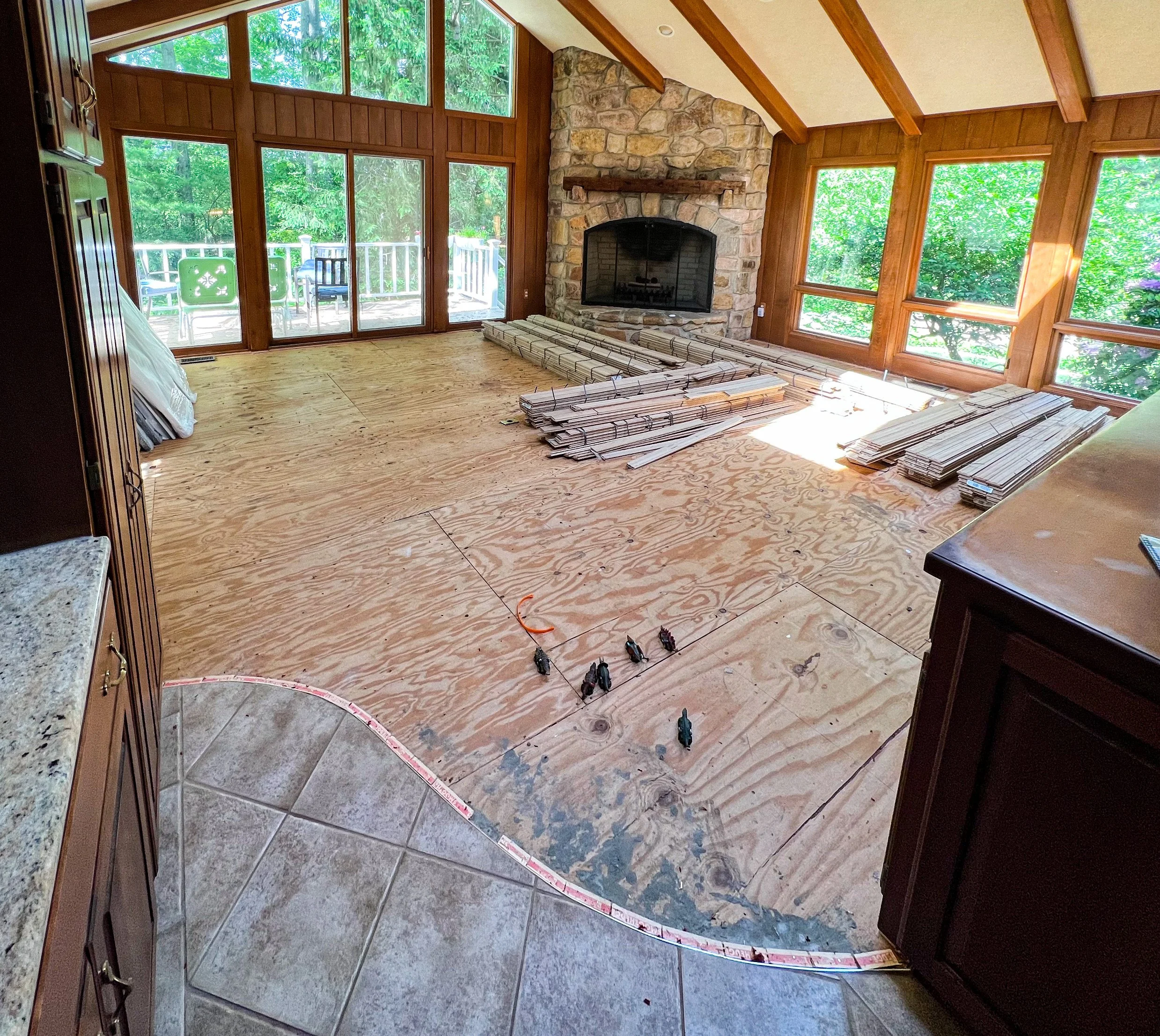 Living room under renovation with unfinished wooden floor, construction materials, and large windows with a view of green trees outside.