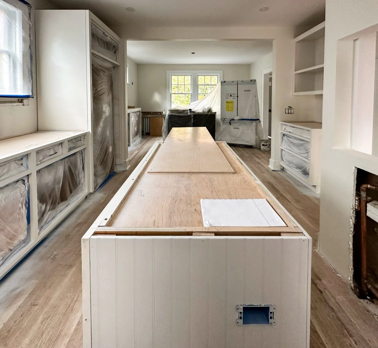 Kitchen undergoing renovation with cabinetry covered in protective plastic and a large center island with unfinished wood surface, surrounded by framed openings for appliances and shelves, with some construction materials visible.