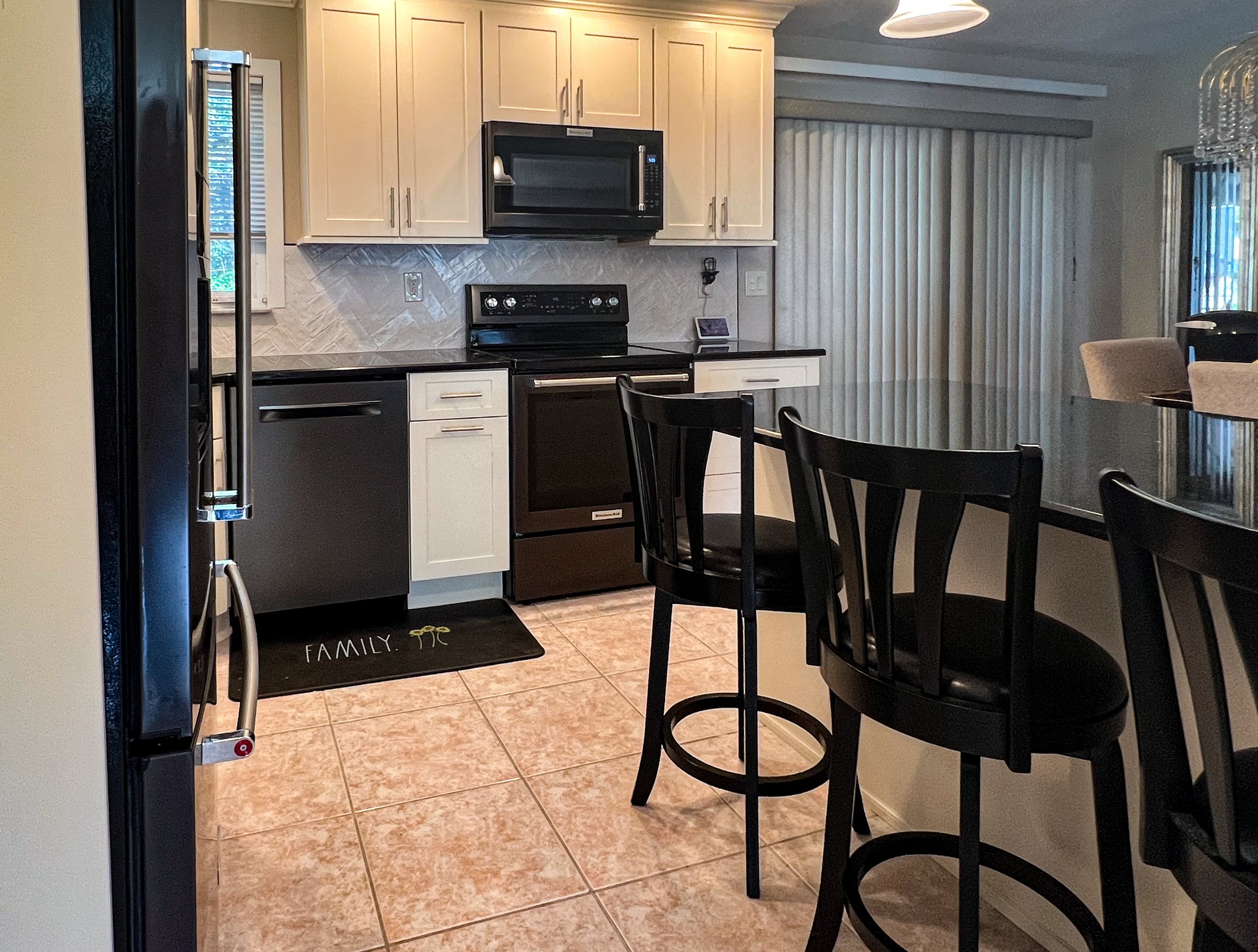 Kitchen with black appliances, white cabinets, and beige tiled floor, featuring a black counter and a modern backsplash. A black barstool is at a countertop, and there is a sliding door with vertical blinds in the background.