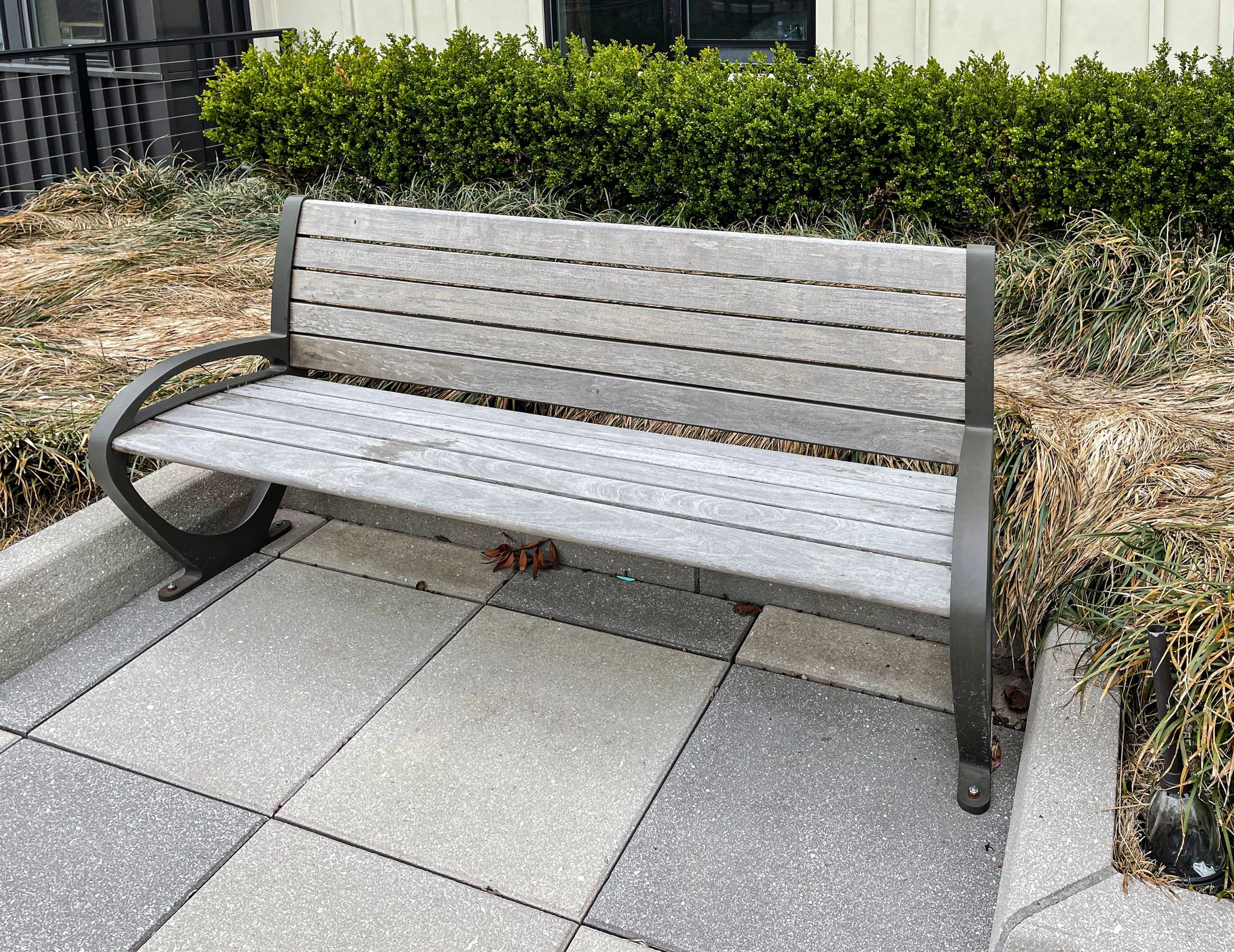 Empty park bench with a metal frame and wooden slats, situated on a concrete sidewalk with a bushy planting area behind it.