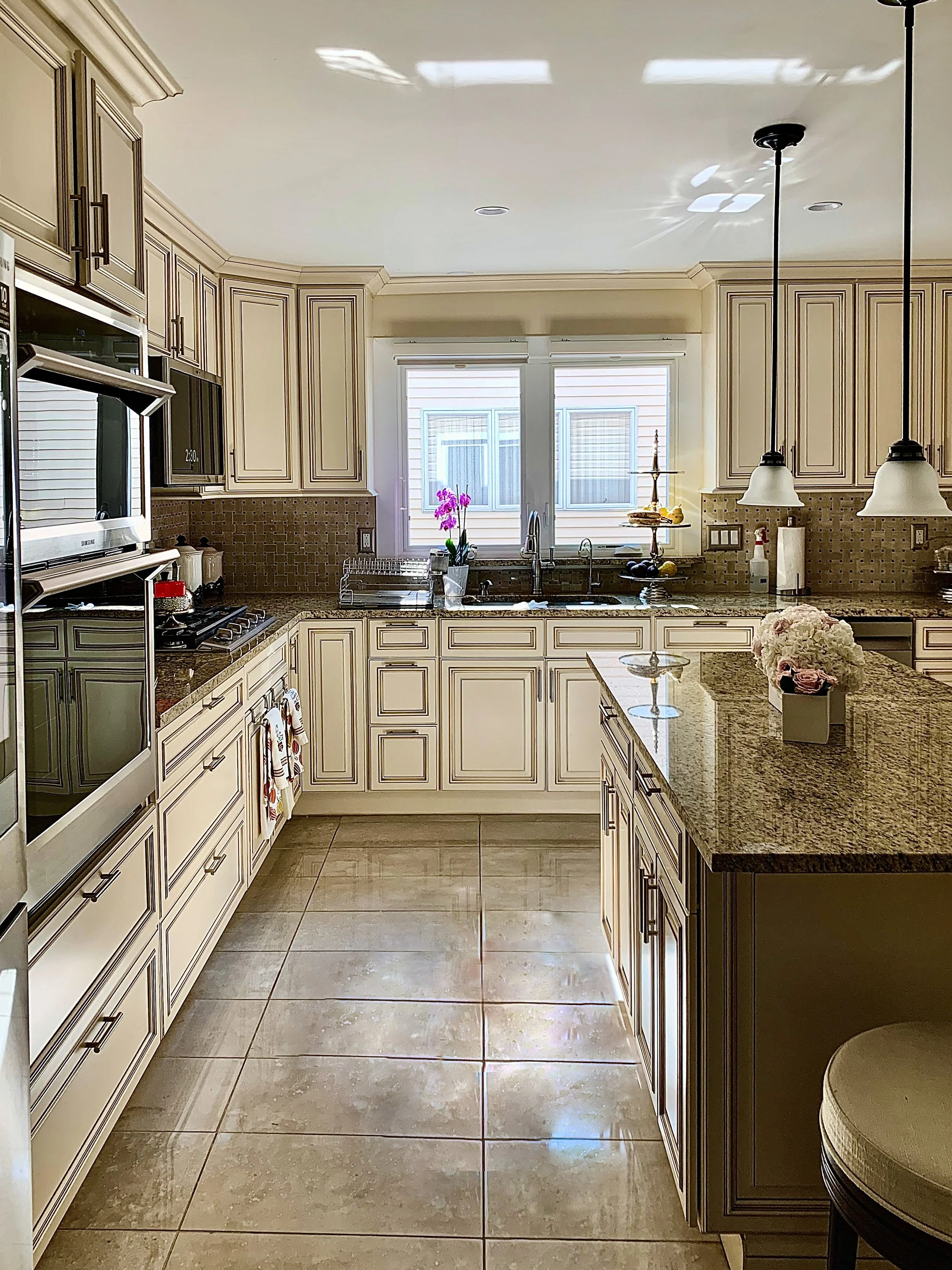 A well-lit kitchen with cream-colored cabinets, granite countertops, and large beige tiled floors. There is a window above the sink with pink flowers, and various kitchen items on the counters. Two pendant lights hang over an island with a flower arr