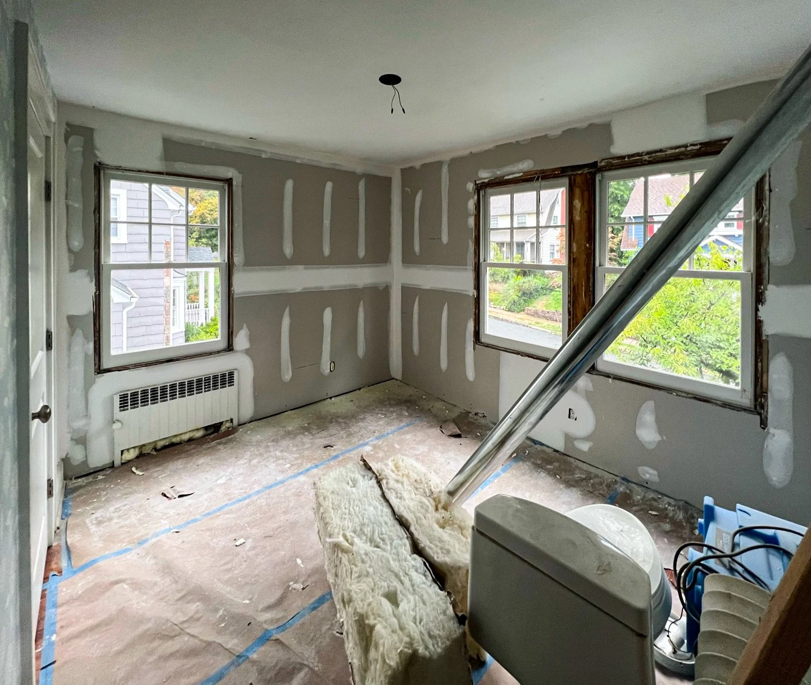 A room under renovation with unfinished drywall on the walls, two windows installed, and construction materials on the floor, including insulation and tools.