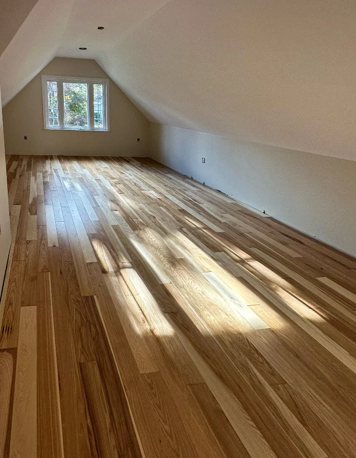 Empty attic room with sloped ceiling, a window showing trees outside, and newly installed wooden flooring.
